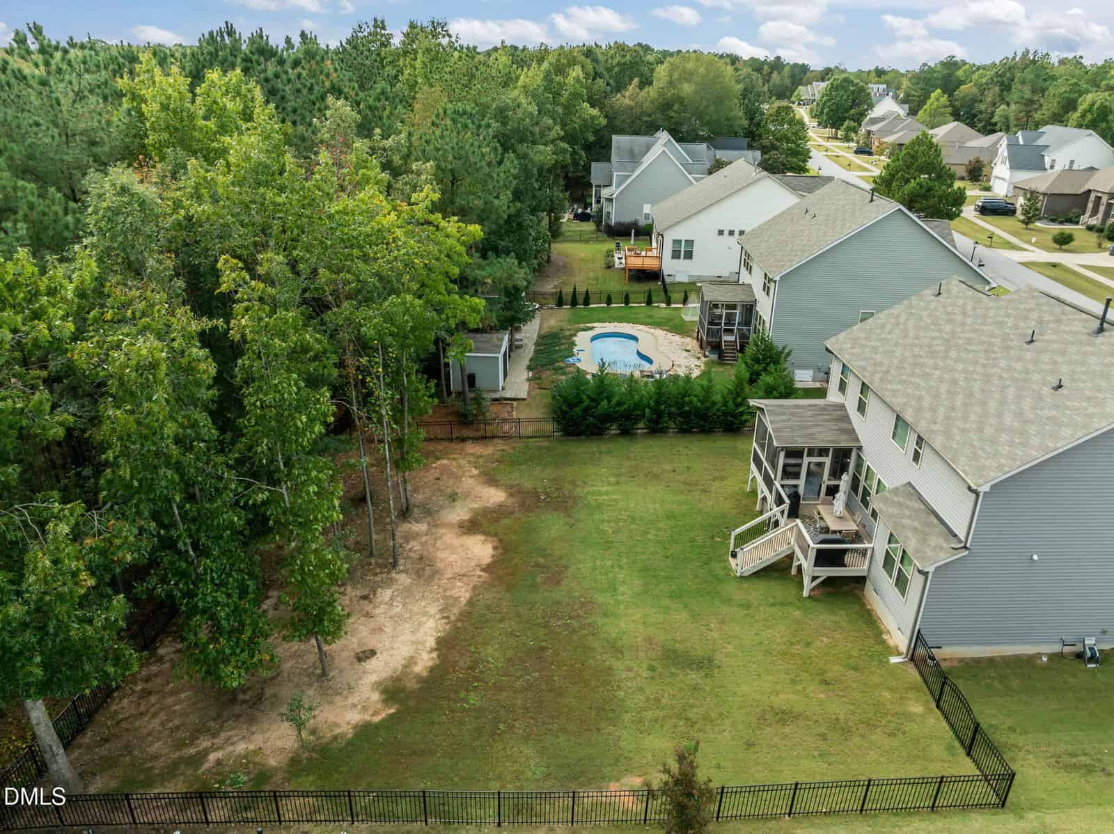 A fenced backyard with patches of grass and dirt, a few trees on the left, and two gray houses; one house has a screened porch and deck, and a swimming pool is visible in a neighbor’s yard.