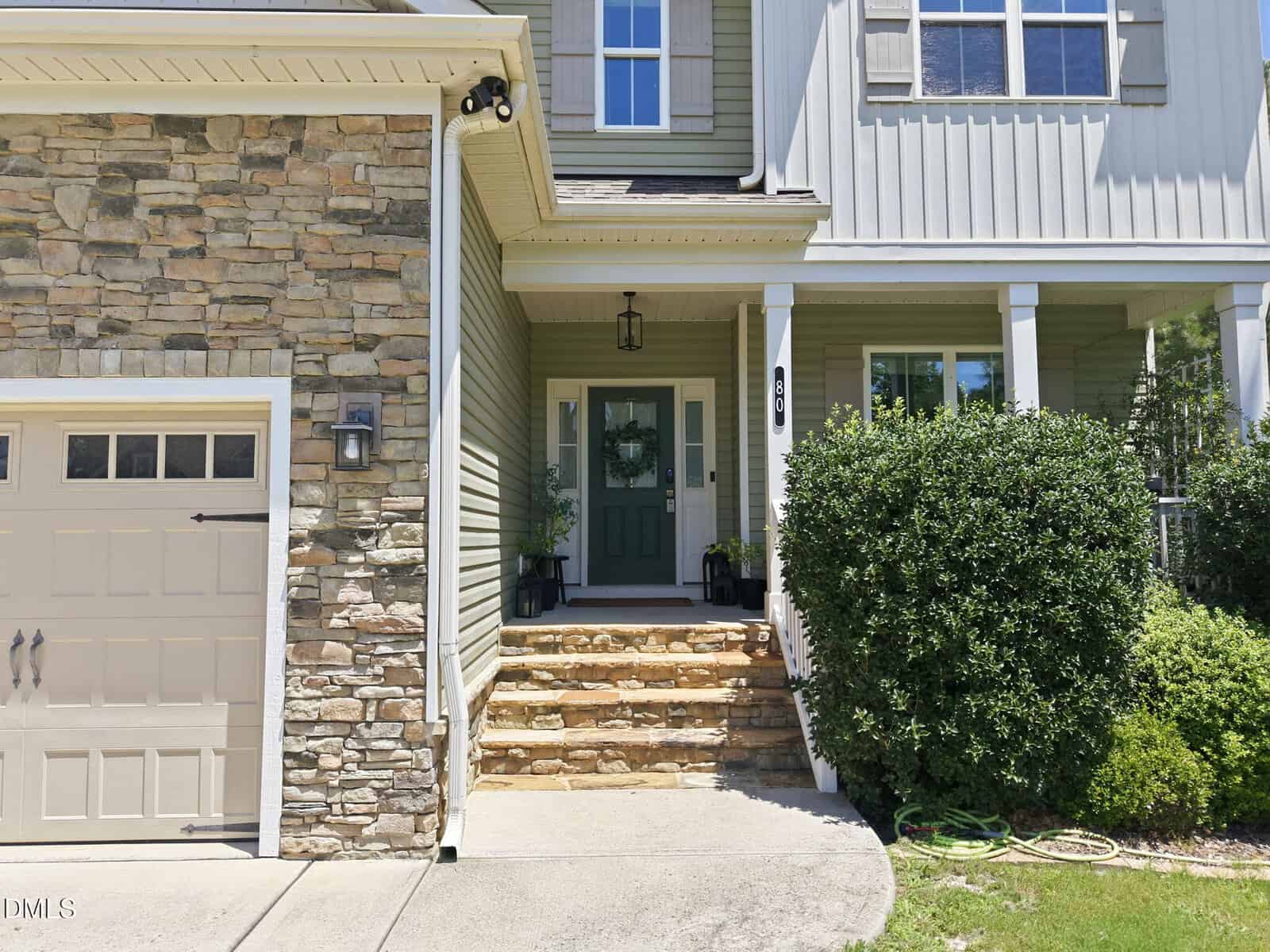 Front entrance of a house with stone and siding exterior, green door with sidelights, three stone steps, white columns, garage to the left, large bush to the right, and a garden hose on the ground.