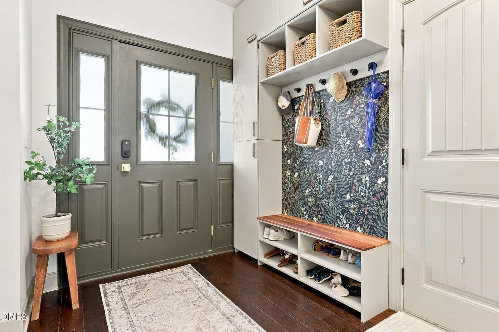 A tidy entryway with a green door, built-in storage with baskets, hooks holding bags and umbrellas, a bench with shoe cubbies, a potted plant on a stool, and a rug on a dark wood floor.