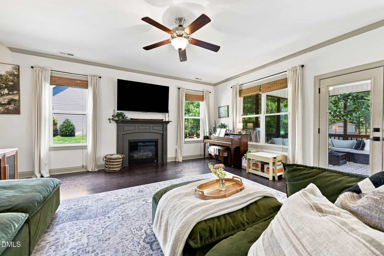 Bright living room with a ceiling fan, green sofa, fireplace, wall-mounted TV, piano, and large windows with white curtains. Hardwood floors and neutral decor create an inviting, cozy atmosphere.