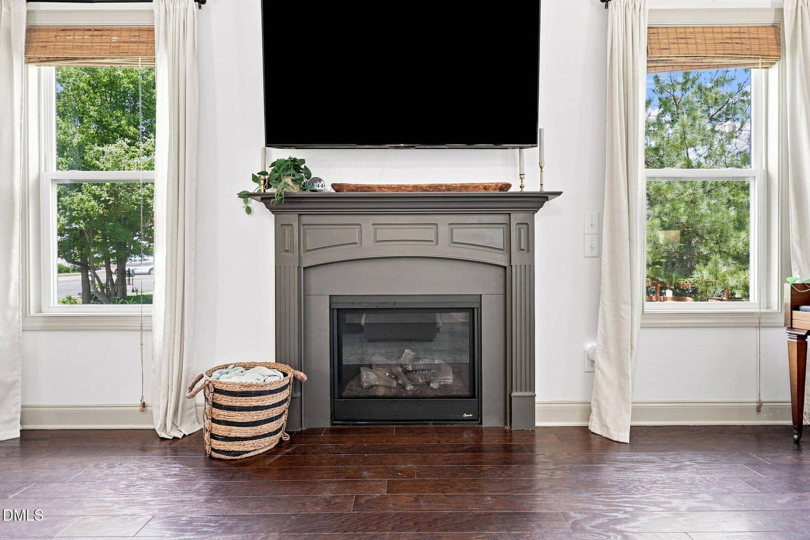A dark wood fireplace sits between two windows with white curtains. A black TV is mounted above the mantel, which holds a plant and a tray. A basket with blankets is on the left, and greenery is visible outside.