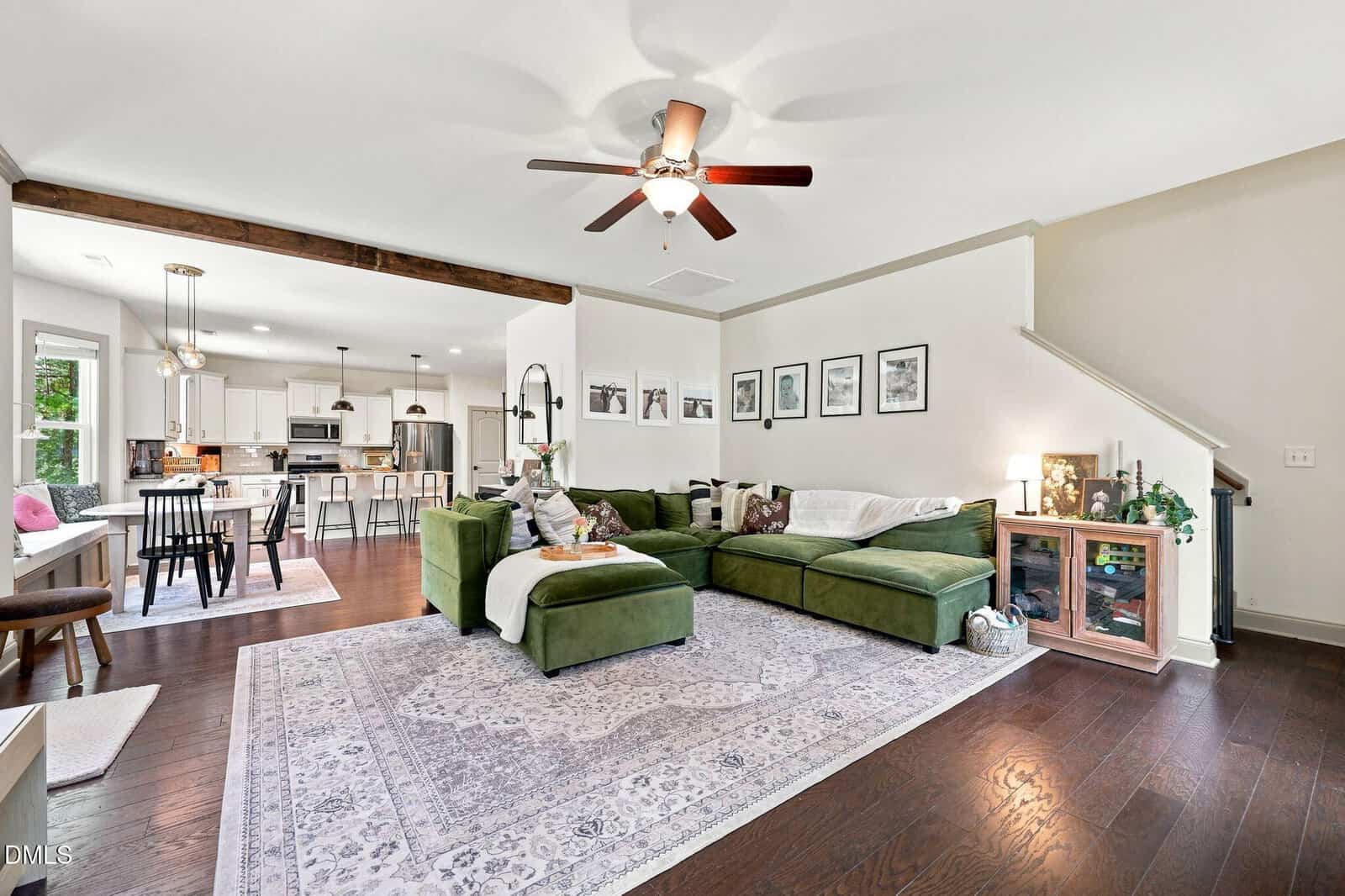 Open-concept living room and kitchen with a green sectional sofa, dining table, modern kitchen, wood floors, ceiling fan, and framed art on the wall. Stairs and a glass cabinet are also visible.