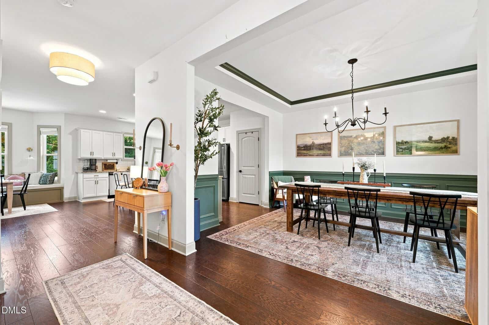 Bright open dining and kitchen area with dark wood floors, green wainscoting, white walls, a long dining table with black chairs, kitchen with white cabinets, wall art, and rugs on the floor.