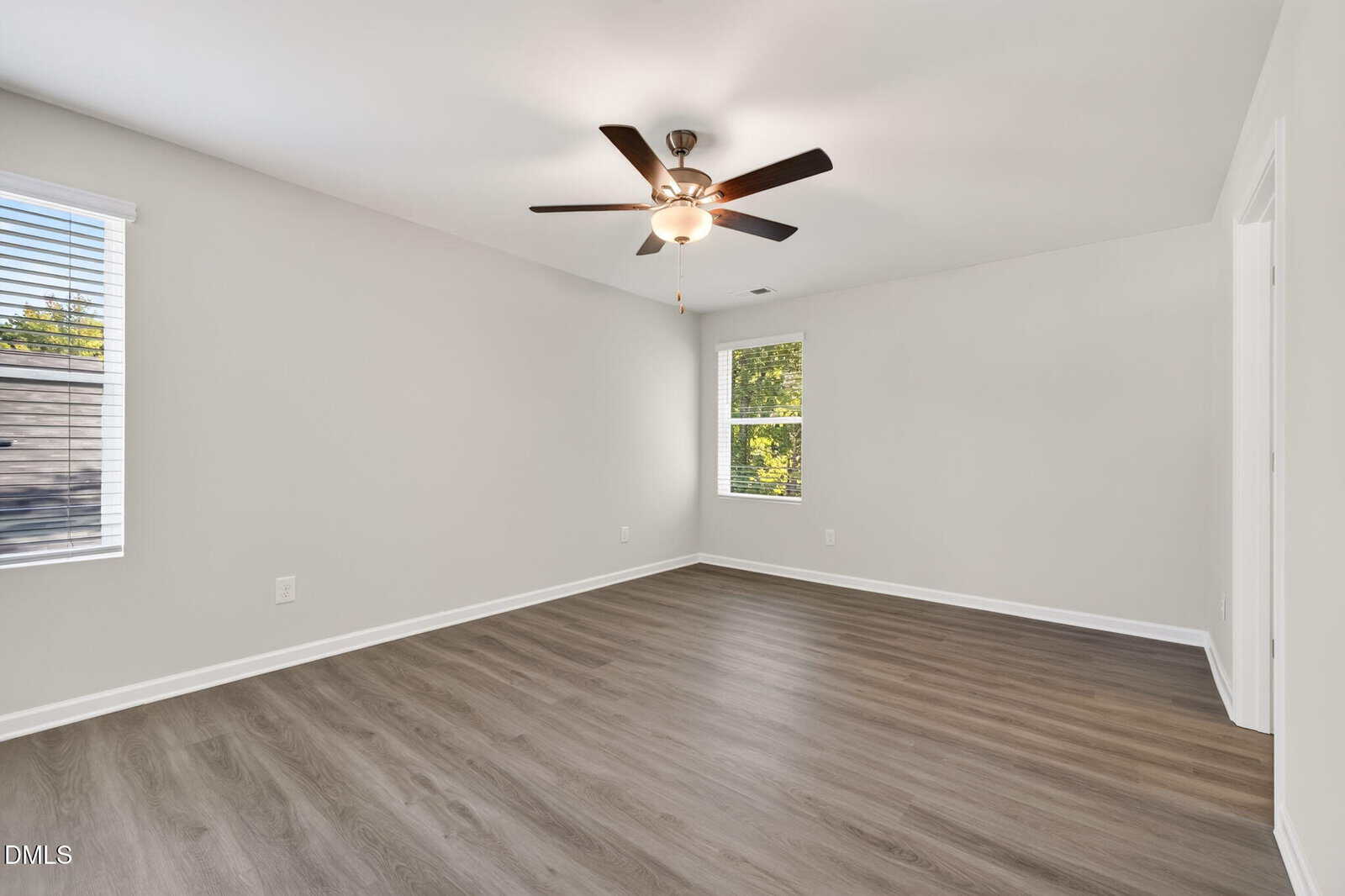 Empty room with light gray walls, wood-style flooring, two windows with blinds, and a ceiling fan with a light. Natural light enters through the windows, creating a bright and spacious atmosphere.