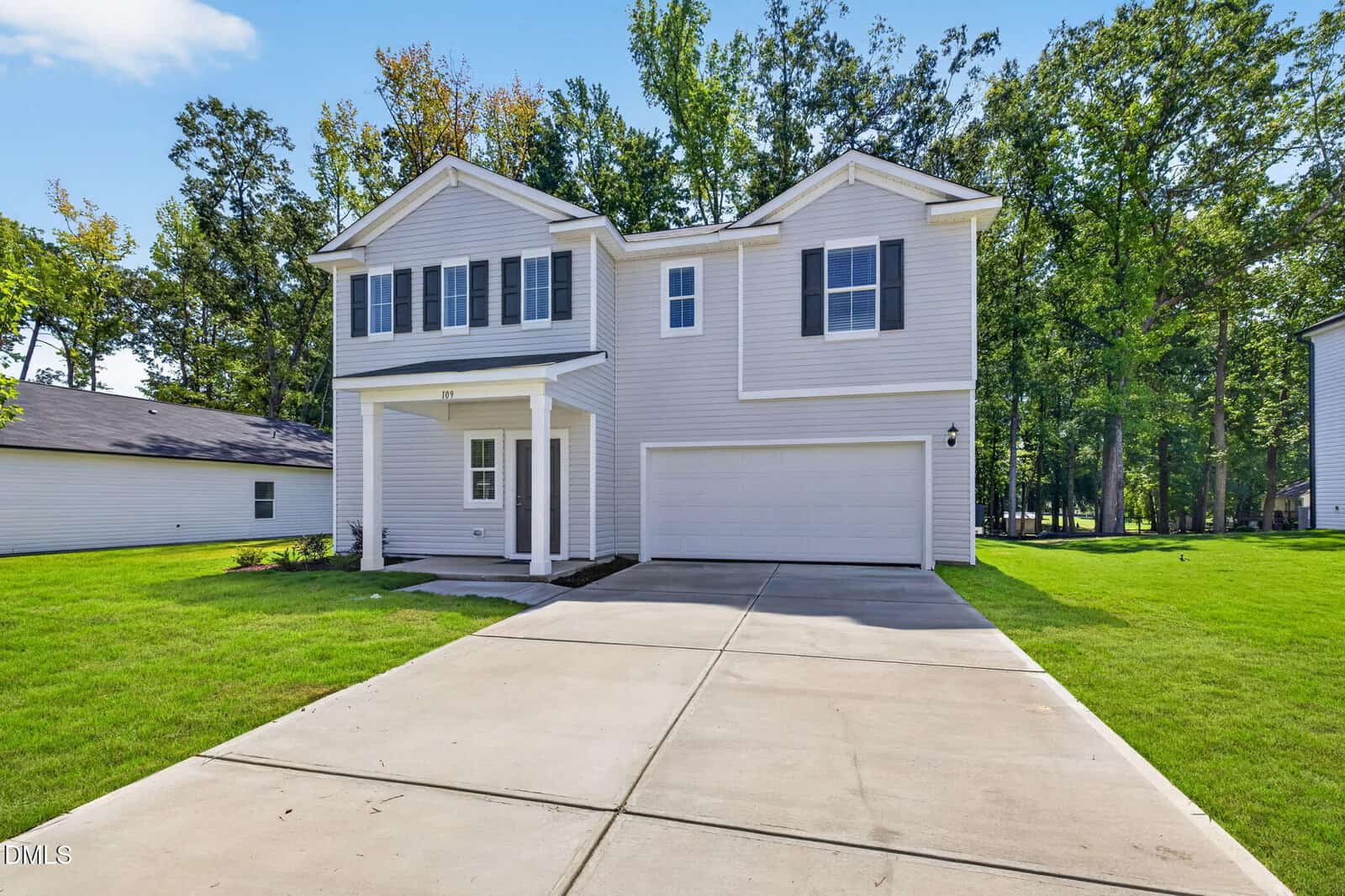 Two-story light gray house with white trim, black shutters, a covered entry, and a two-car garage. The house is surrounded by green grass, trees, and a clear blue sky.