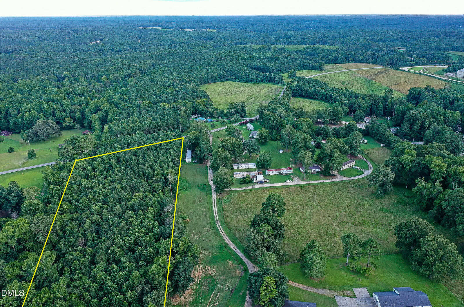 Aerial view of a rural landscape with dense forest outlined in yellow, bordered by open fields, scattered houses, winding roads, and extensive wooded areas in the background.