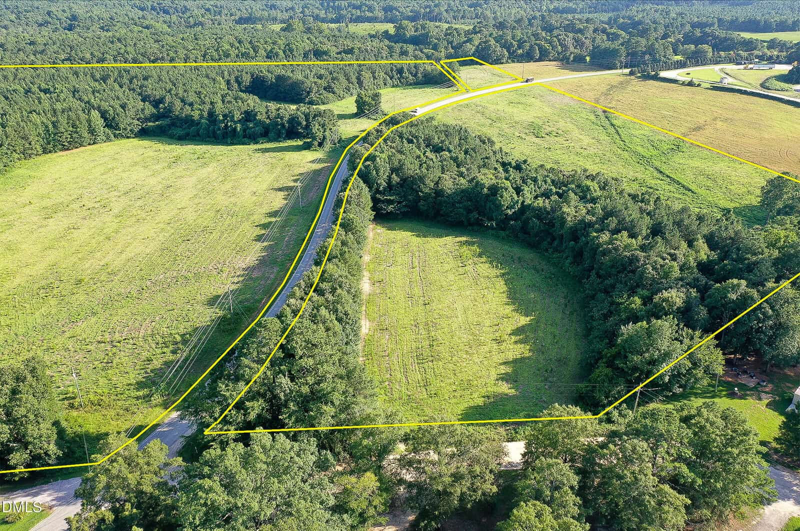 Aerial view of a large, grassy property bordered by trees, with yellow lines outlining its boundaries. A paved road curves through the landscape. Surrounding areas include forests and open fields.
