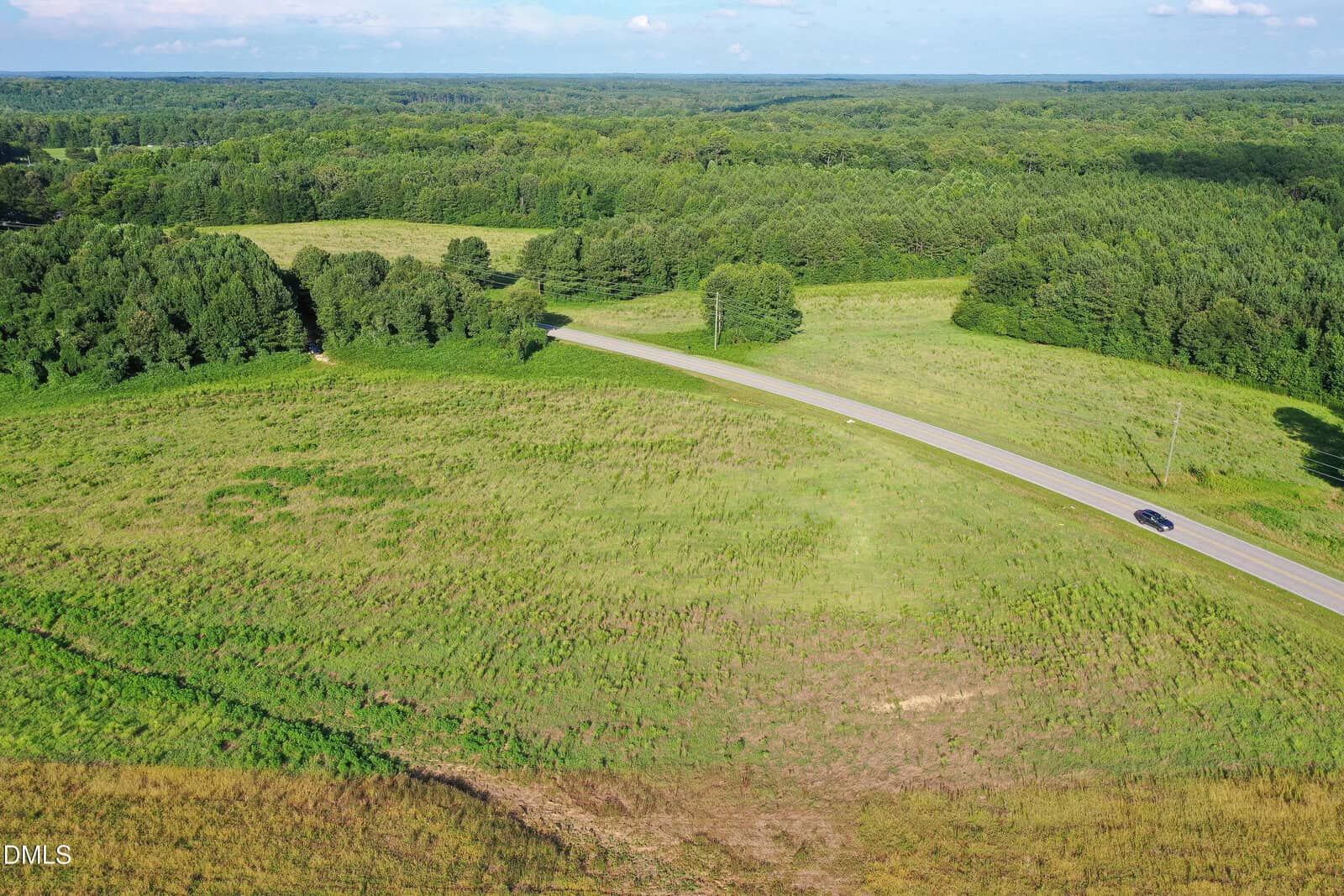 A paved road runs through grassy, open fields bordered by dense green trees under a blue sky with clouds. A black car drives along the road. The landscape is mostly rural and undeveloped.