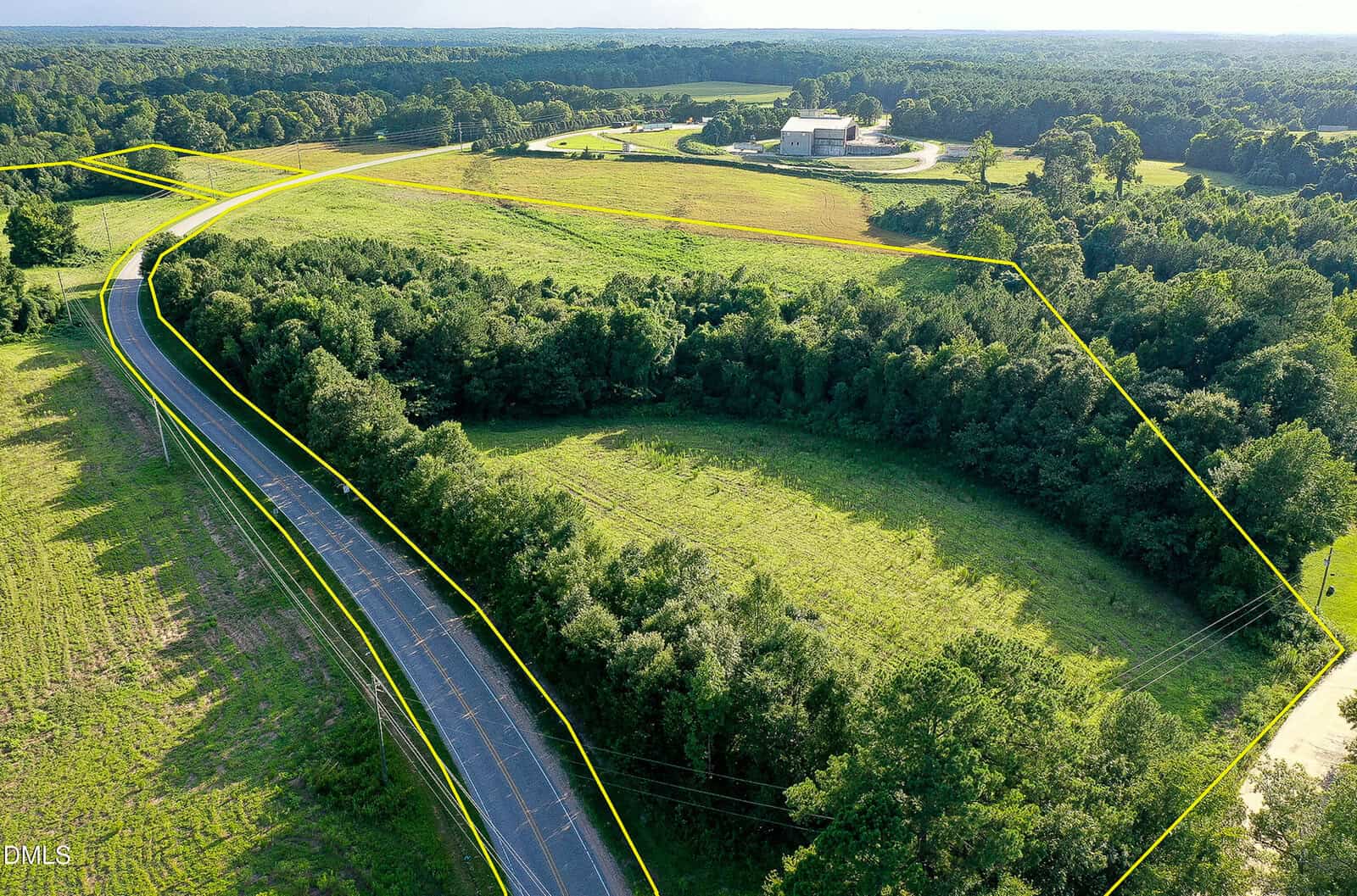Aerial view of a rural property with fields and trees bordered by a yellow outline, showing a curved road on the left and a large building with a driveway in the background.