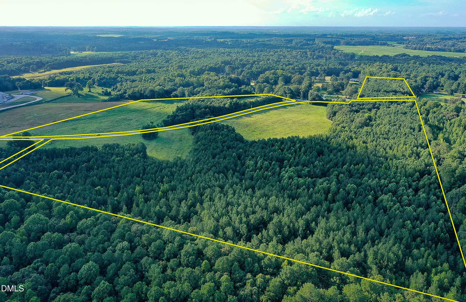 Aerial view of a large, forested property with open fields, outlined by yellow lines to show boundaries. The landscape includes dense woods, grassy areas, and a distant road on the left side.