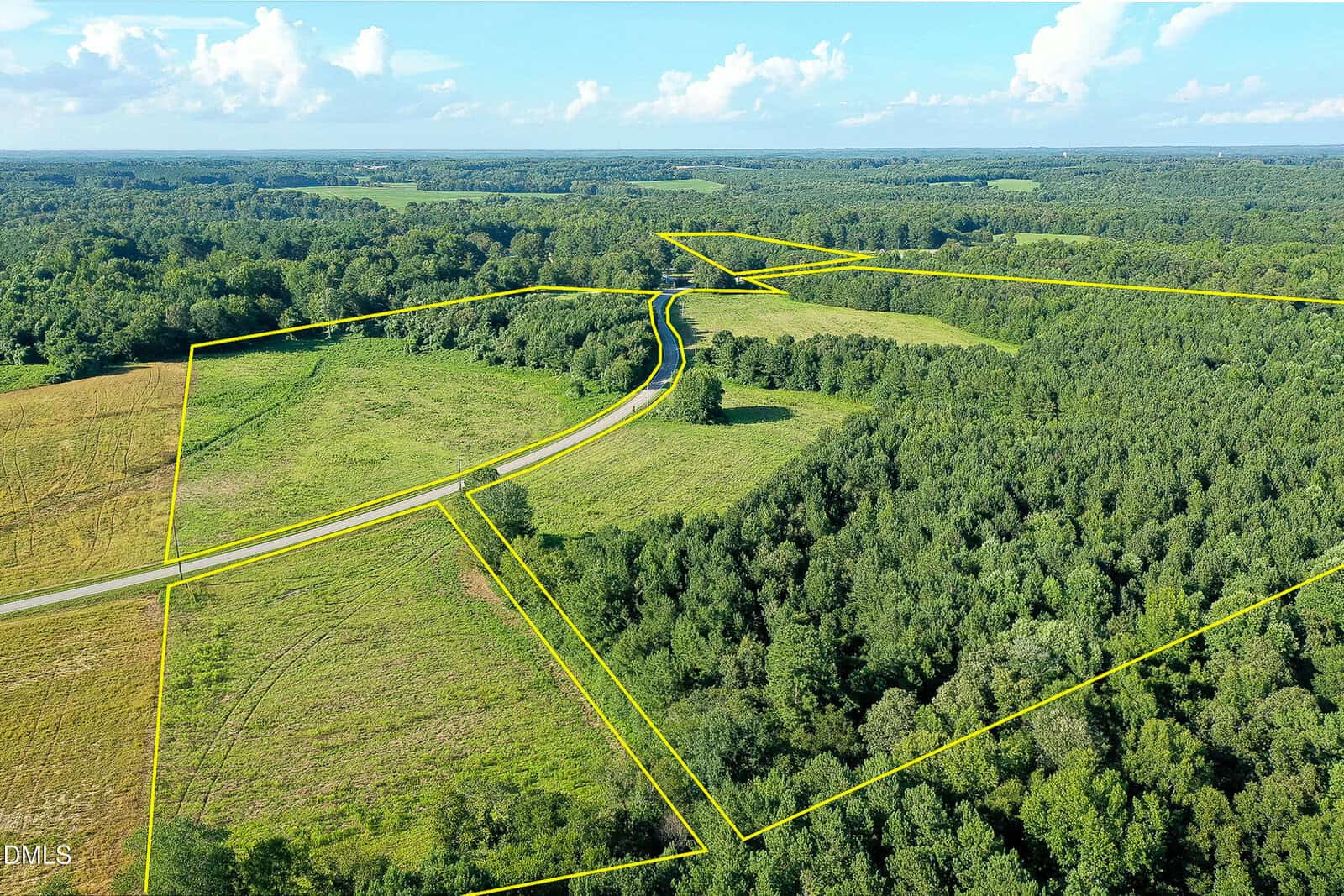 Aerial view of a rural landscape with a road running through fields and forested areas; yellow lines outline multiple plots of land under a blue sky with scattered clouds.