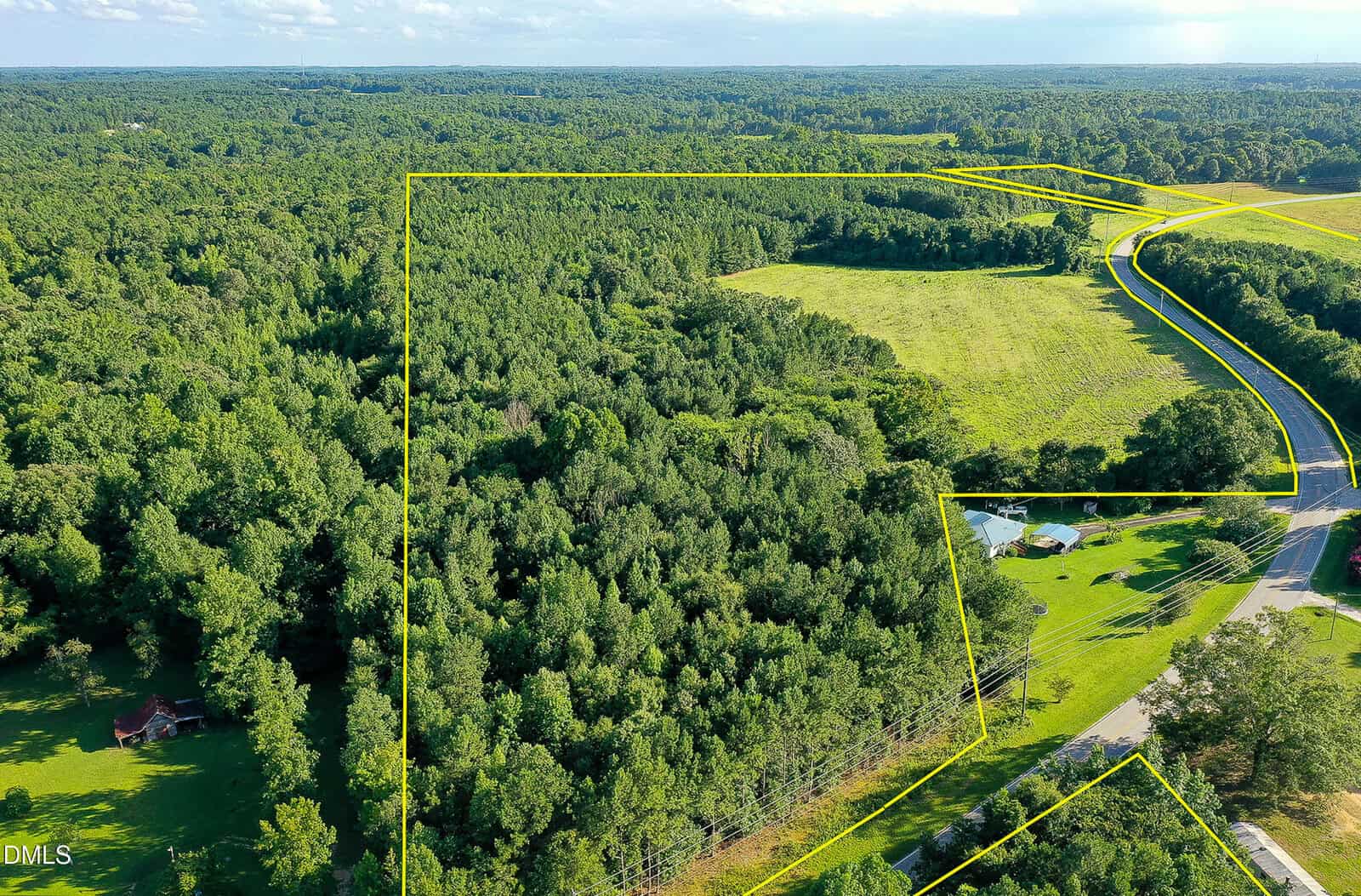 A large plot of wooded land bordered by yellow lines, with open fields and a few scattered houses nearby. A road runs along the edge of the property, and the area is surrounded by dense forest.