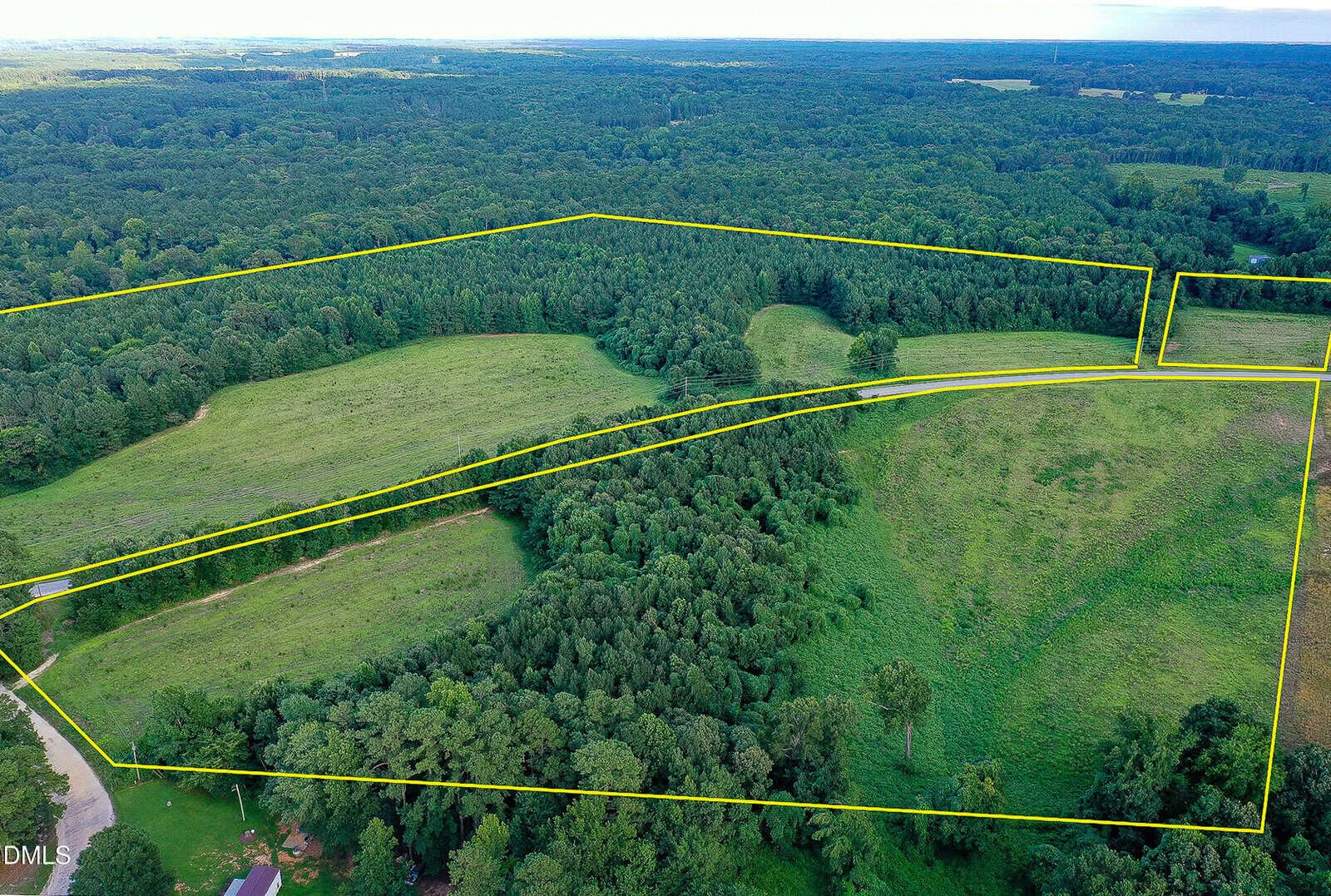 Aerial view of a large green landscape with forests and open fields, outlined by yellow boundary lines to indicate property borders; distant trees and a blue sky are visible in the background.