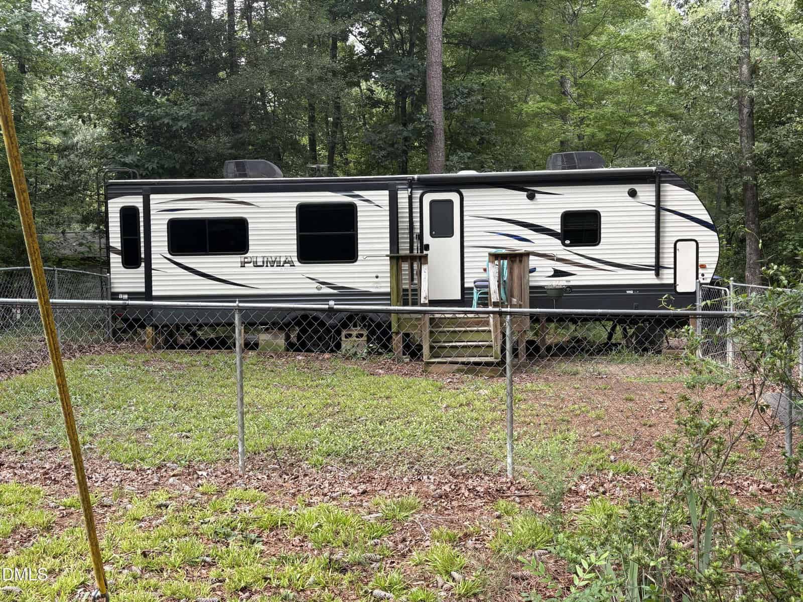 A white and black travel trailer sits behind a chain-link fence in a grassy, wooded yard at 108 Poteau Drive in Louisburg. There are wooden steps leading to the door, and trees surround the area.