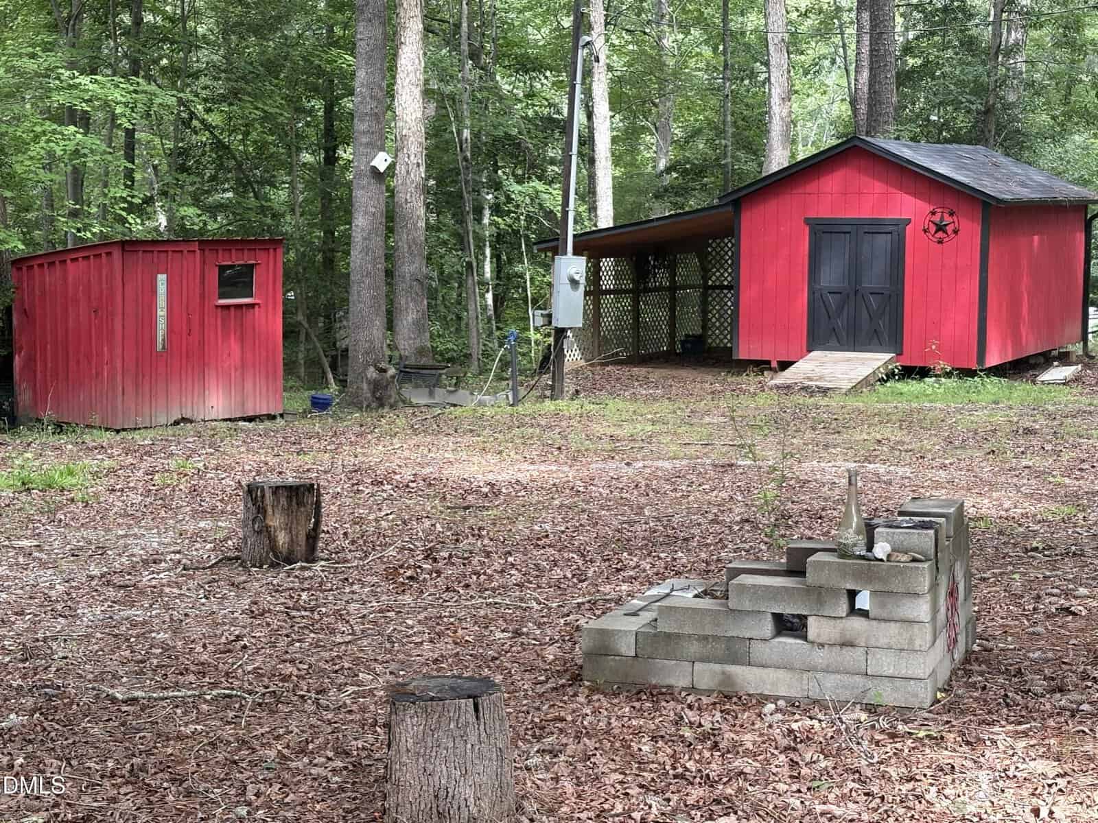 Two red wooden sheds sit among trees in a wooded area. In the foreground, there are tree stumps and a small structure made of concrete blocks. The ground is covered with dry leaves and dirt.