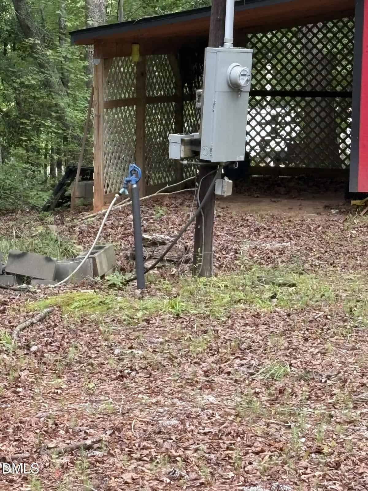 A utility meter and electrical box are mounted on a wooden pole near a building with lattice siding. The area is covered with dry leaves and some grass; trees and woods are in the background.
