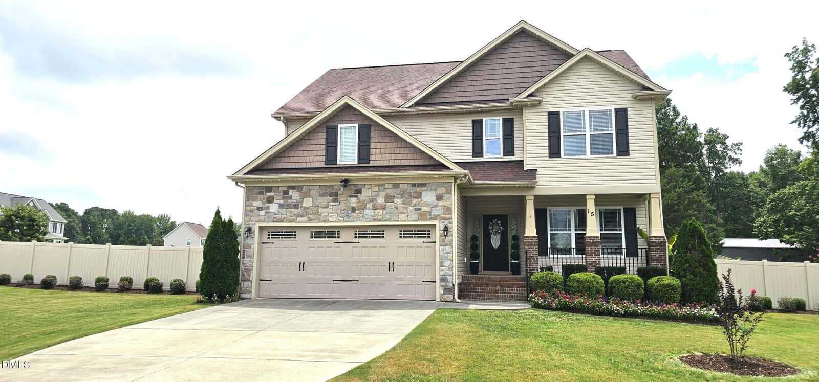 Two-story suburban house at 15 Brushwood Court, Youngsville, with tan siding, stone accents, a double garage, and a manicured front yard featuring shrubs and a small flower bed. The driveway leads to the garage, and a white fence encloses the backyard.