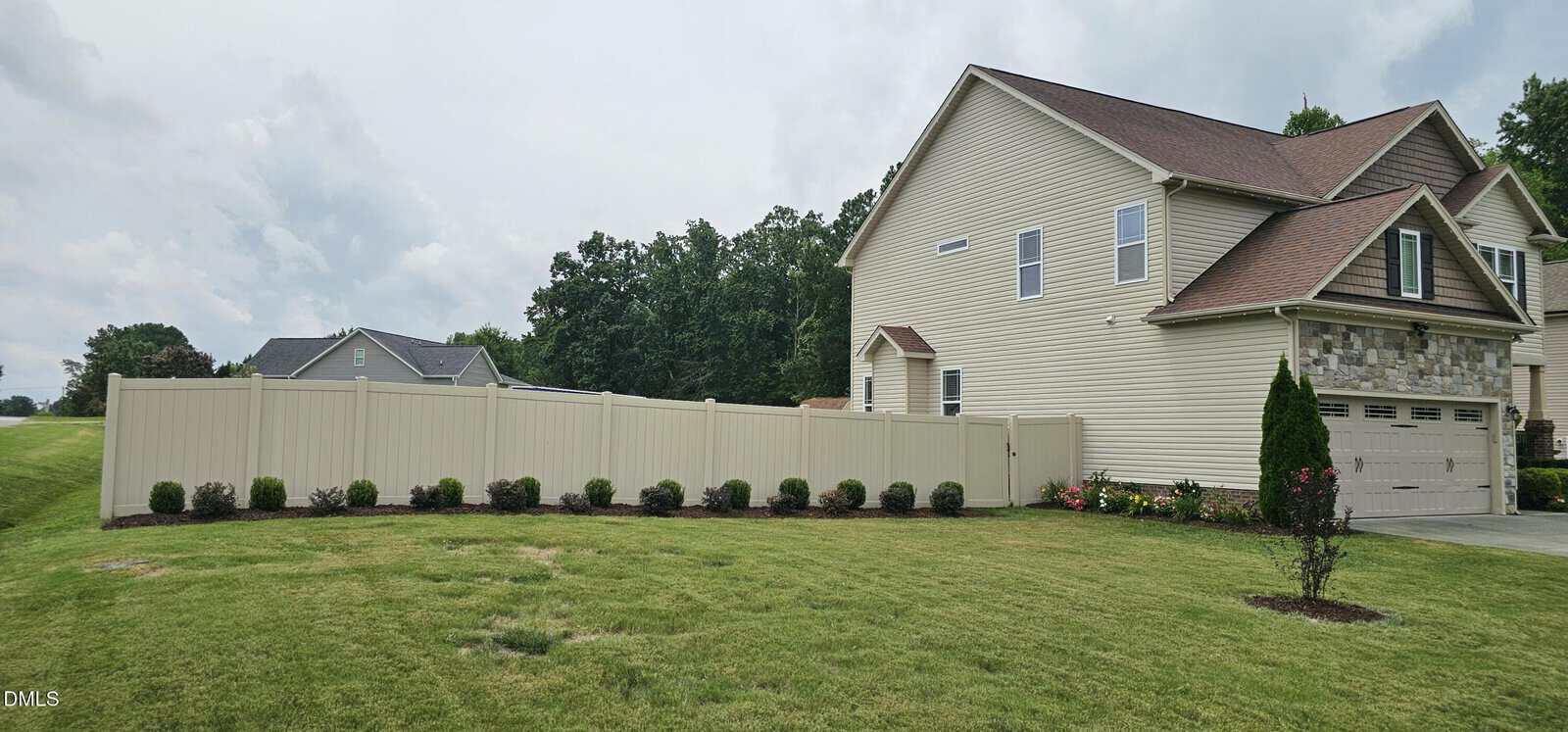 A beige two-story suburban house with a fenced backyard, neatly trimmed lawn, small shrubs along the fence, and a paved driveway. The sky is cloudy and trees line the background.