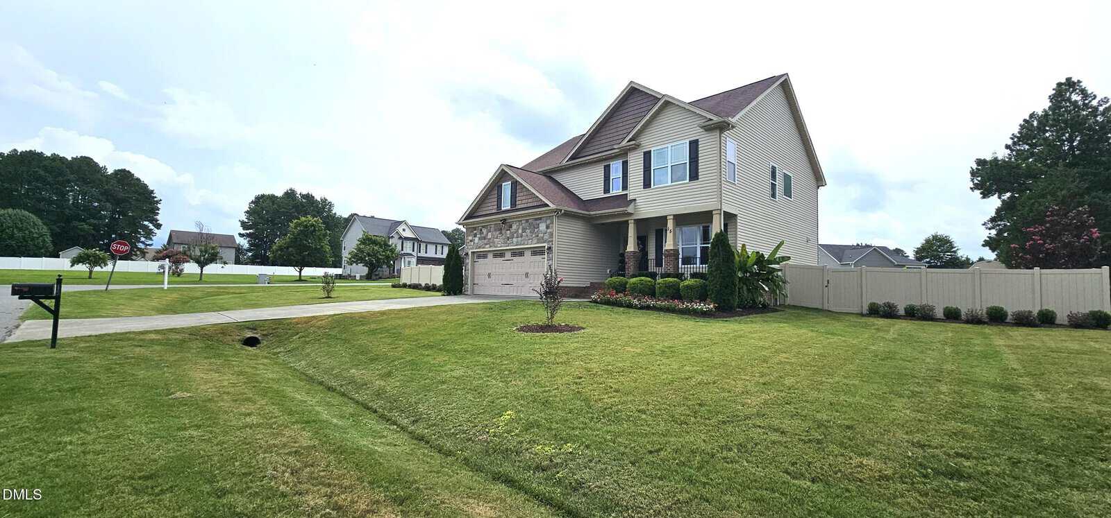 Two-story beige house with a three-car garage, front porch, and landscaped yard on a corner lot. The house has green grass, shrubs, and a fenced backyard. Neighboring homes and a street are visible in the background.