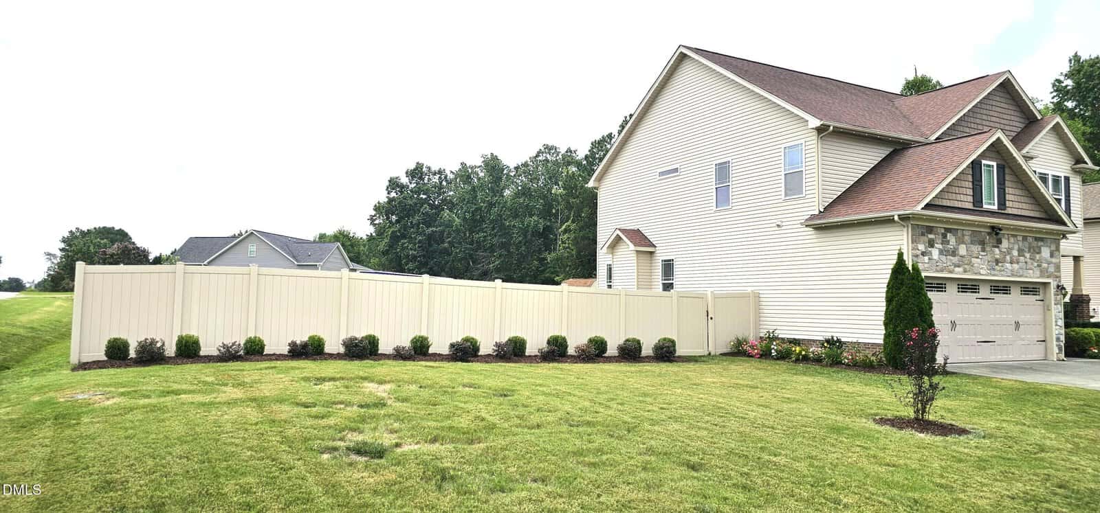 A beige two-story suburban house with a stone accent, attached garage, light-colored privacy fence, small shrubs along the fence, and well-maintained lawn. Other houses and trees are visible in the background.
