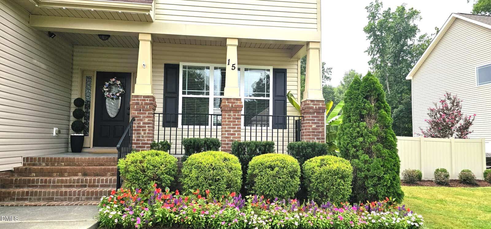 A beige two-story house with a brick porch, black front door, large front window, black railing, and neatly trimmed bushes and flowers in the front yard. A neighboring house and trees are visible in the background.