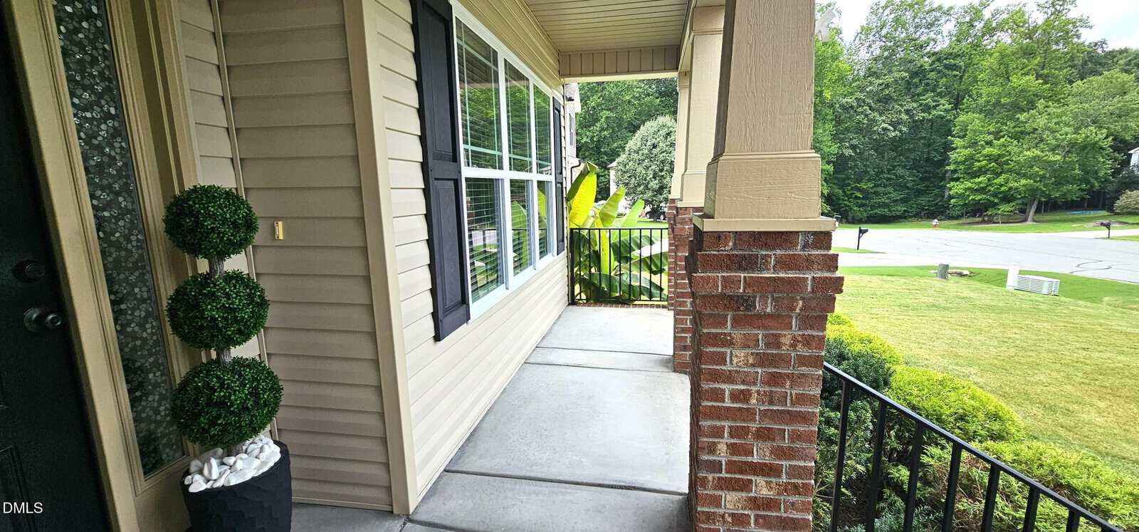 A covered front porch with beige siding, brick columns, a black metal railing, and two potted topiary plants, overlooking a grassy yard and a quiet suburban street with trees in the background.
