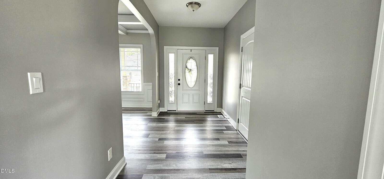 A bright entryway with gray walls, wood-look flooring, a white front door with glass panels, side windows, a ceiling light, and an arched doorway leading to another room with a window.