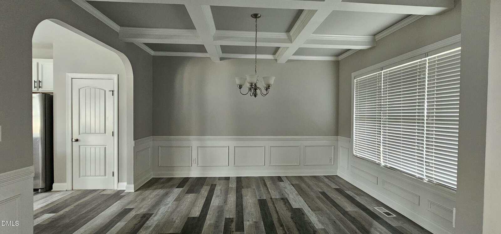 A spacious, empty dining room with gray walls, white trim, a coffered ceiling, chandelier, large window with blinds, and wood-look flooring in varying shades of gray and brown.