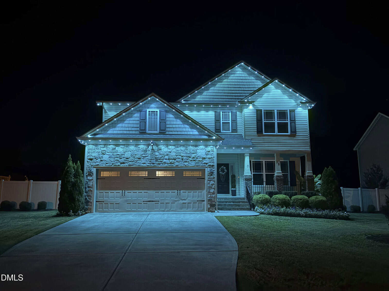 Two-story house with stone and siding exterior, illuminated by blue lights along the roofline at night. The driveway is empty, and the yard is neatly landscaped with shrubs and grass.