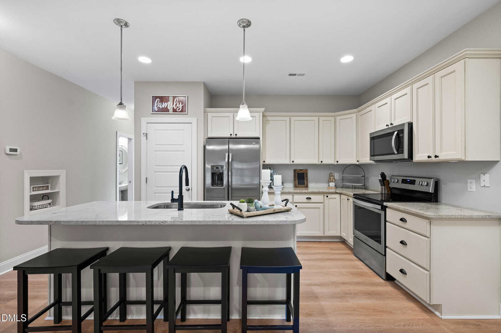 Modern kitchen with white cabinets, stainless steel appliances, an island with a sink, and four black stools. Pendant lights hang above, and a “Laundry” sign is on the wall near a white door.