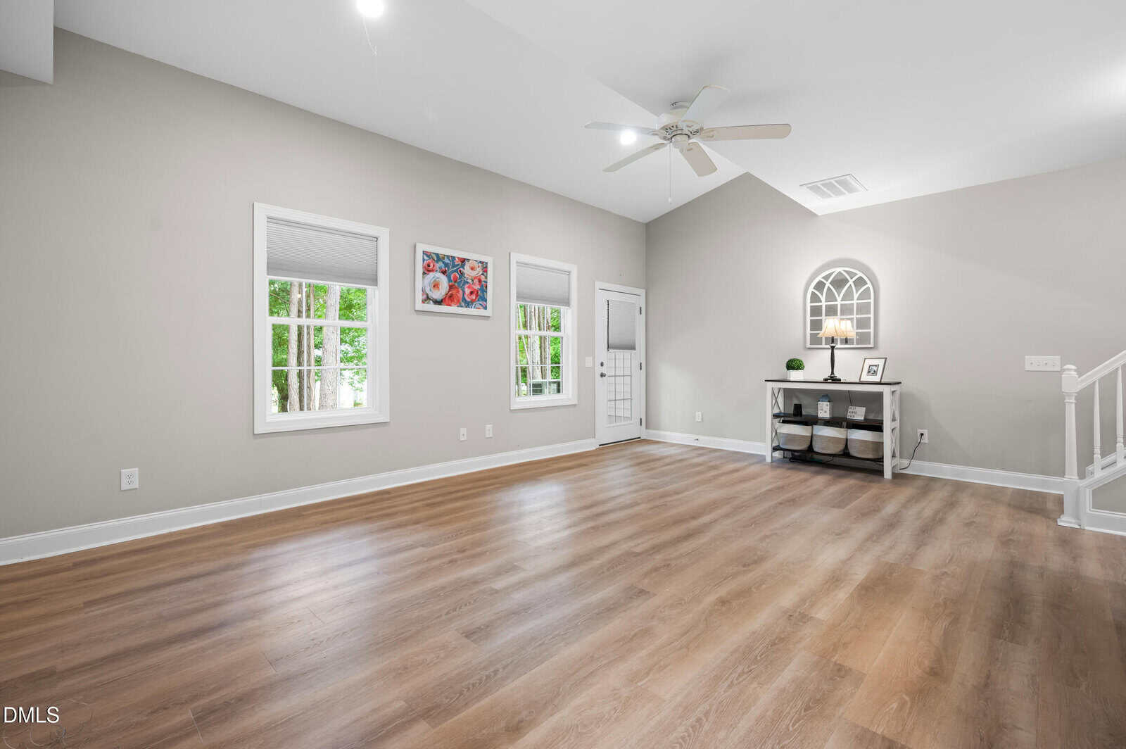 Living room with light wood flooring, pale gray walls, three windows, and a white ceiling fan. A small table with storage baskets, decor, and framed photos is against the wall near a door and staircase.