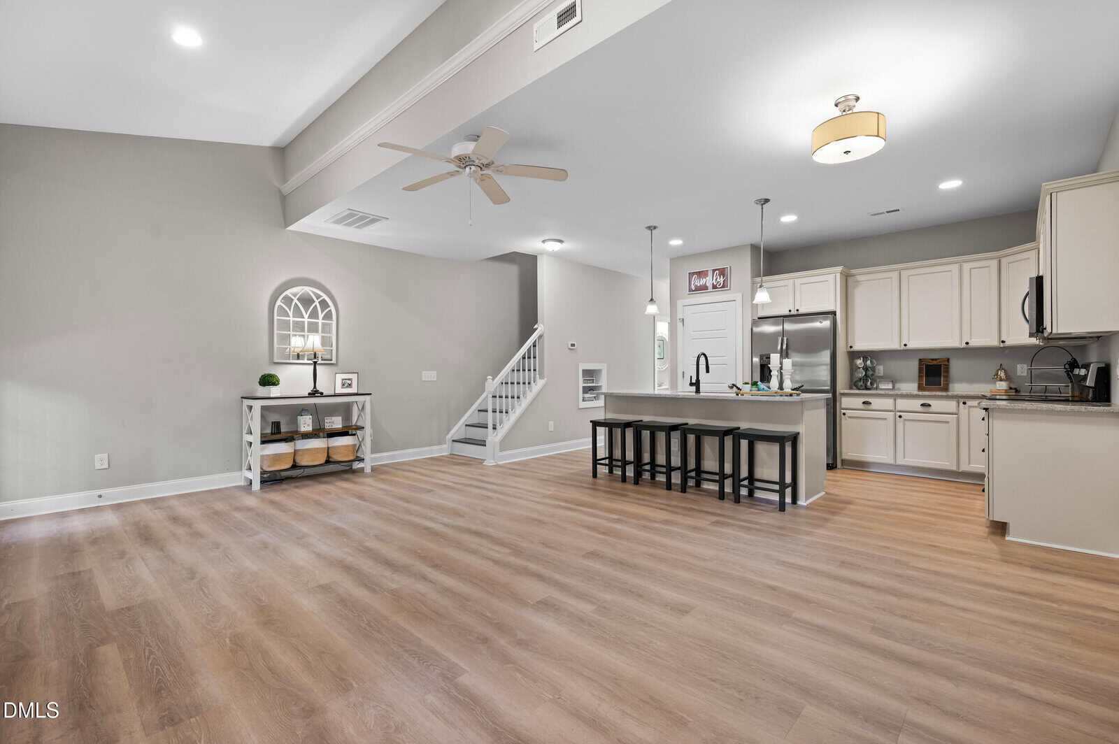 Open-concept kitchen and living area with light wood flooring, neutral walls, white cabinetry, stainless steel appliances, a center island with stools, ceiling fan, and stairs leading to the upper level.