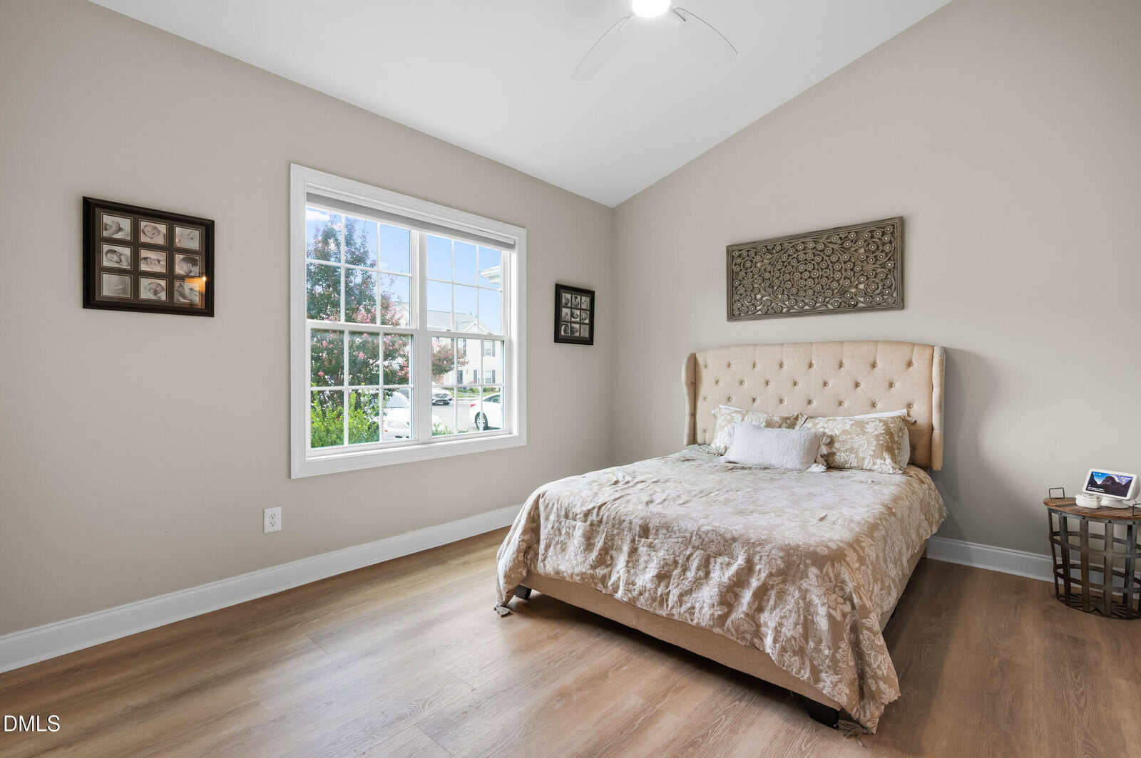 A bedroom with a beige upholstered bed, neutral bedding, a window with outdoor view, wood flooring, and framed wall art. A small side table with a digital device is next to the bed. Walls are painted a light color.