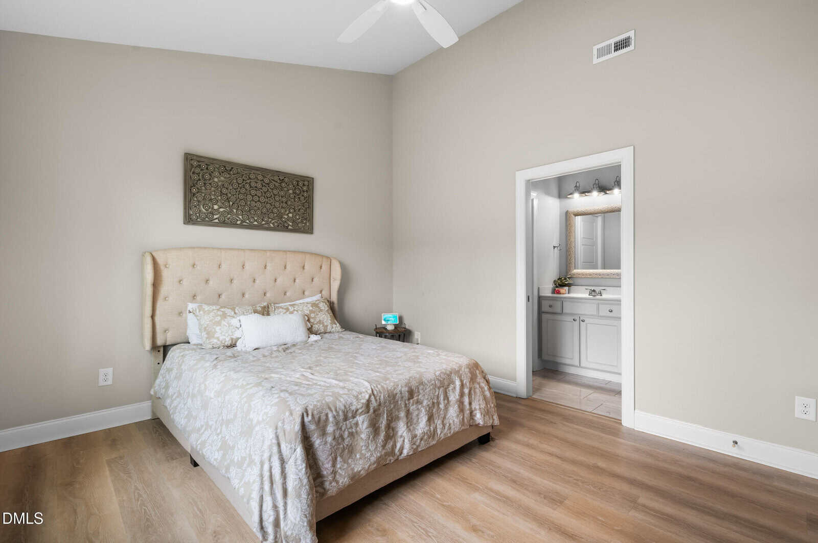 Bright bedroom with a beige upholstered bed, patterned bedding, and decorative pillows. Wooden floor, neutral walls, wall art above bed, and an open doorway leading to a bathroom with a large mirror.