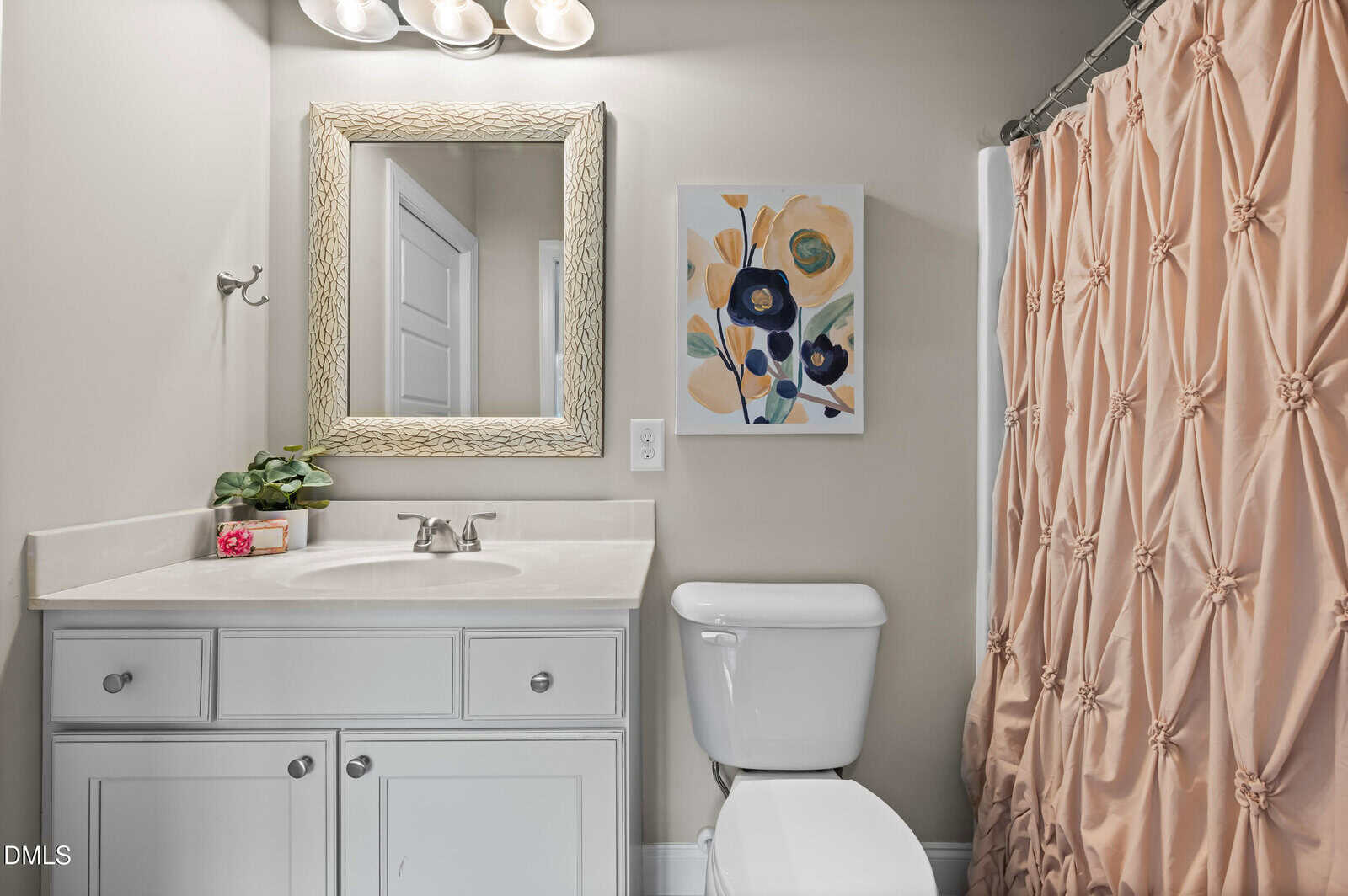 A bathroom with a white vanity, sink, and toilet. A framed mirror and flower artwork hang above. The shower has a pink ruched curtain, and a small plant sits on the counter.