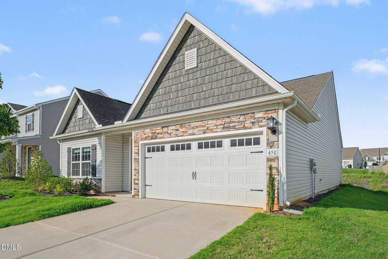 A single-story house with gray siding, stone accents, and a double garage. The driveway leads to a closed garage door with decorative windows. The lawn is green, and neighboring houses are visible in the background.