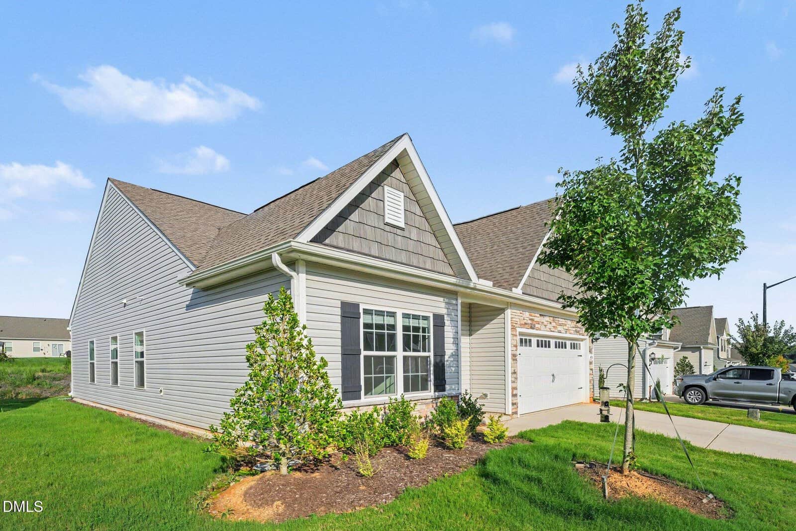 A single-story suburban house with gray siding, a gable roof, attached two-car garage, trimmed shrubs, young trees, and a manicured green lawn under a clear blue sky.