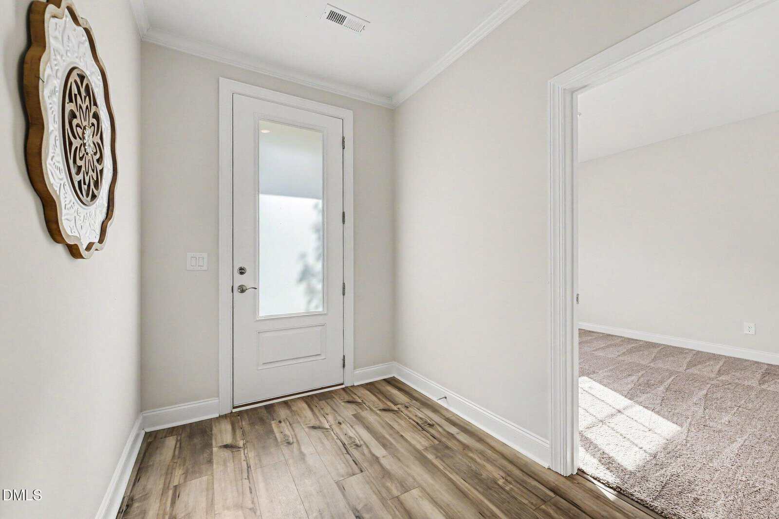 A small hallway with wood flooring, beige walls, a white-framed glass door, a decorative wall piece, and an open doorway leading to a carpeted room with sunlight coming through a window.