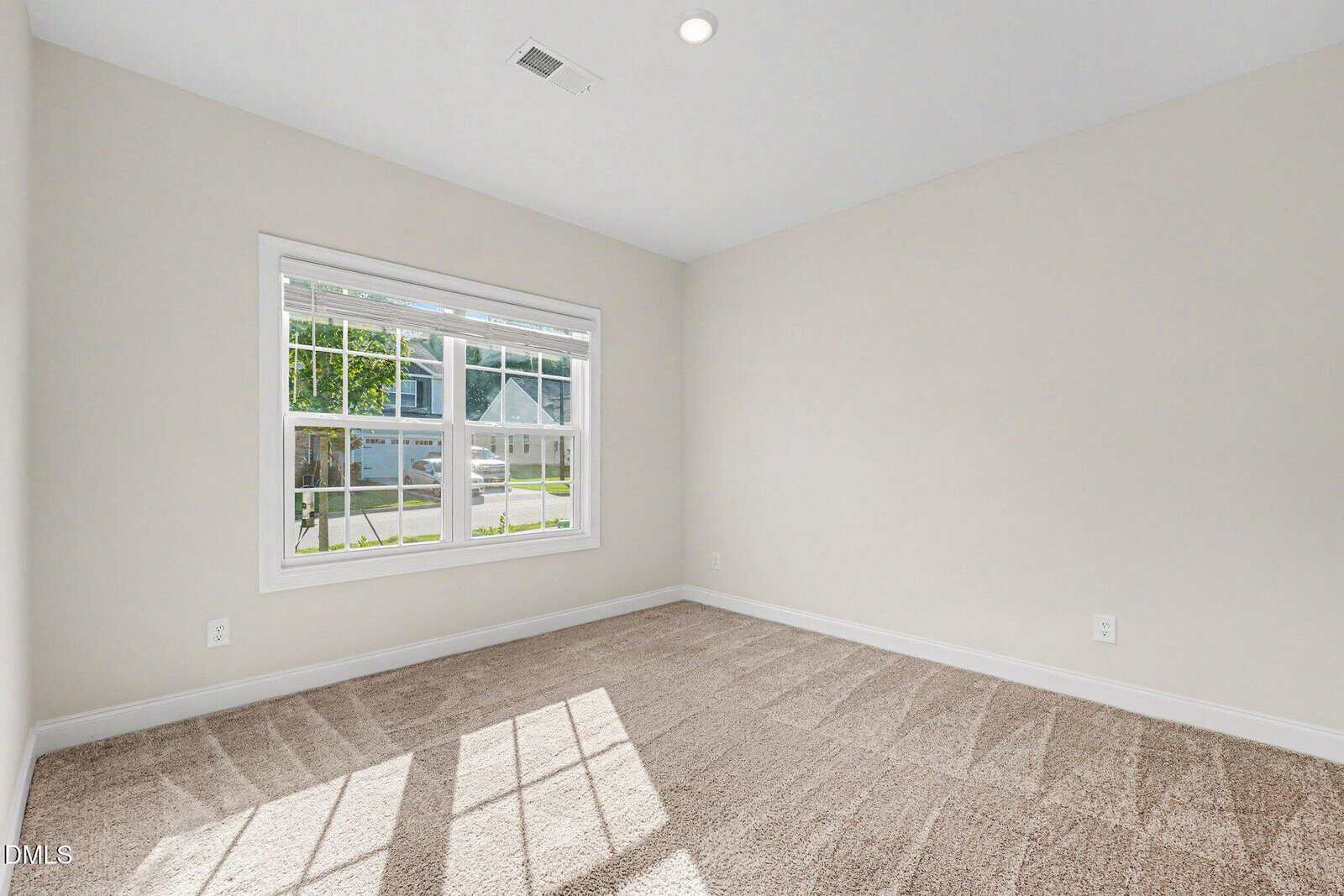 Empty room with beige carpet, light-colored walls, a white ceiling, a central ceiling light, and a large window letting in sunlight. The window overlooks a suburban street with a house and green lawn visible outside.