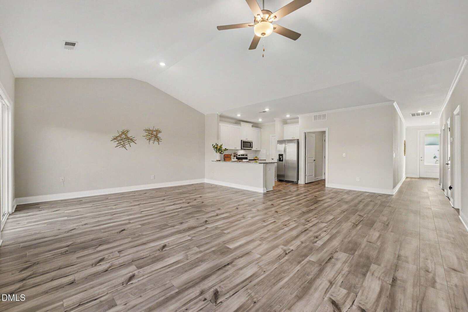 Spacious open-concept living area with light gray wood flooring, white walls, ceiling fan, modern kitchen with stainless steel appliances, and recessed lighting. Artwork decorates one wall. Doorways lead to other rooms.