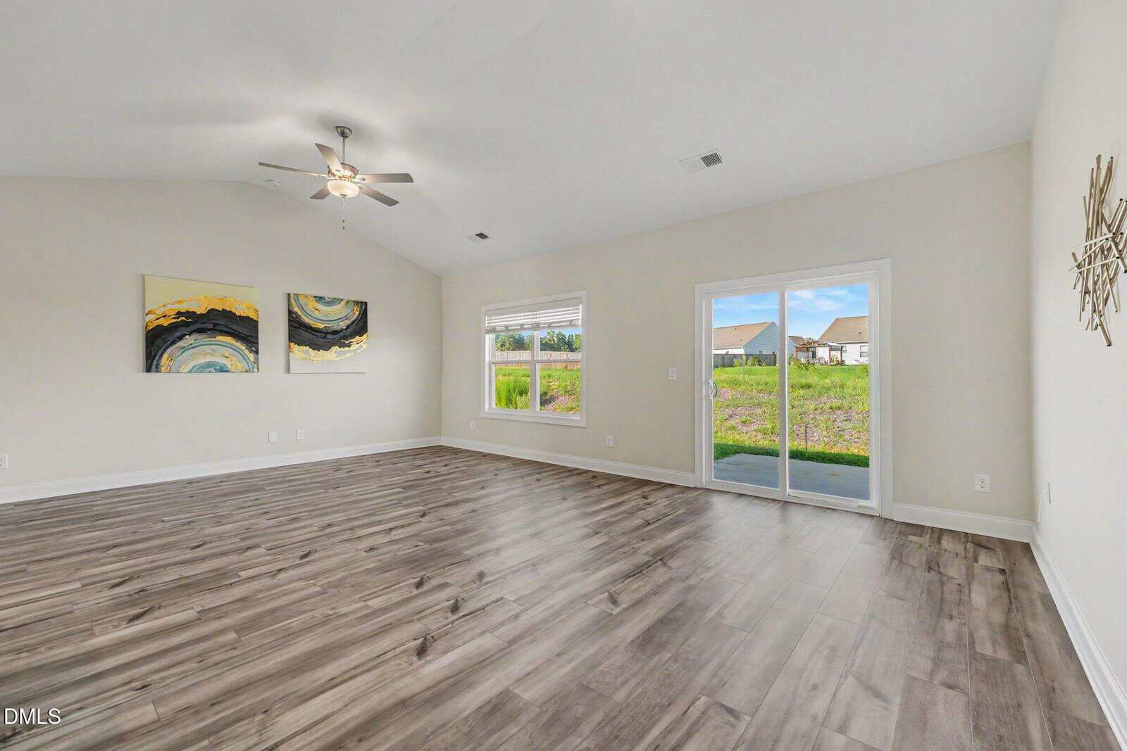 Spacious, empty room with wood flooring, white walls, large window, sliding glass door to a patio, ceiling fan with lights, and modern abstract wall art.