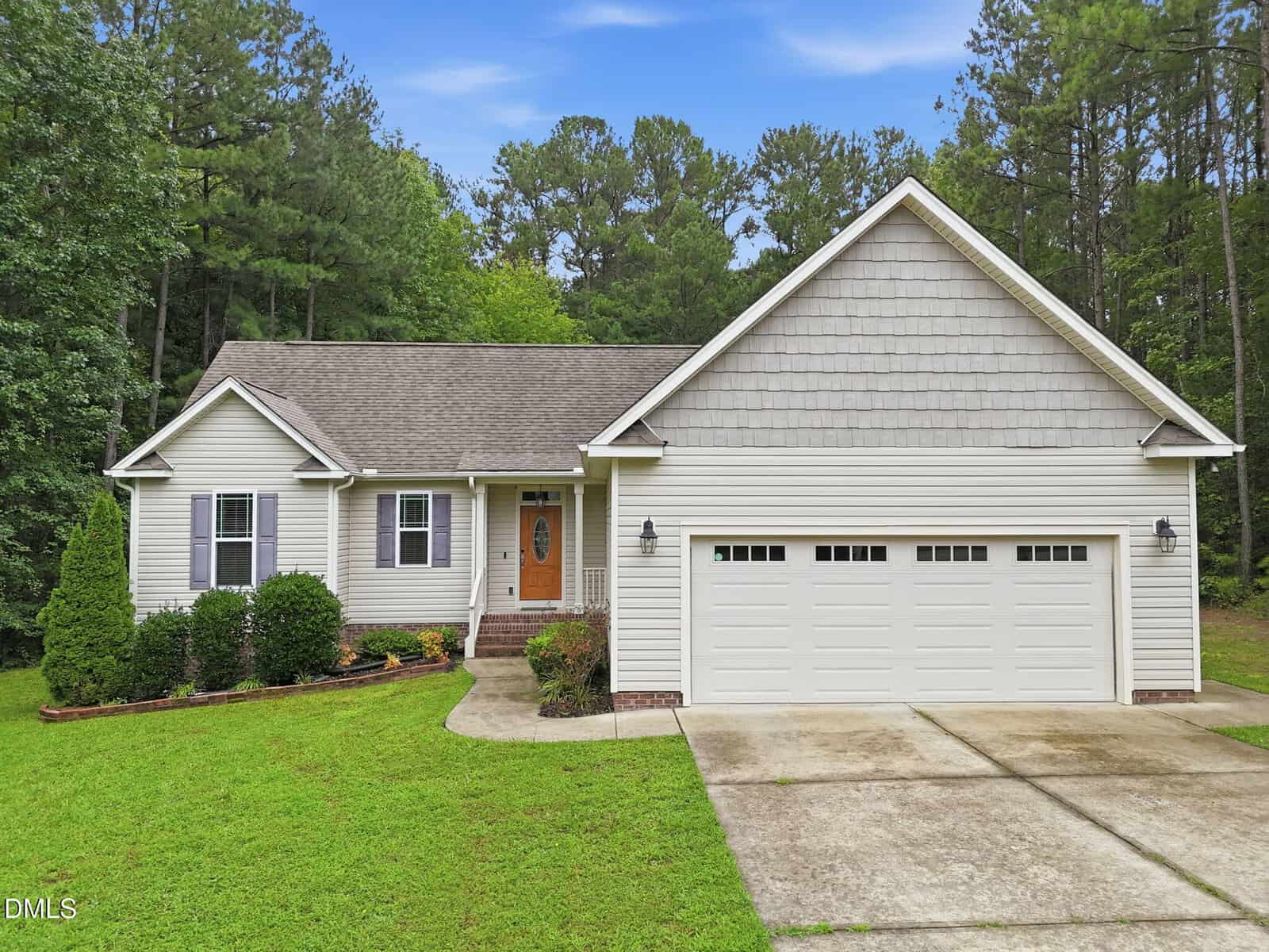 Charming single-story beige house at 85 Copper Creek Drive, Youngsville, featuring a welcoming front porch, gabled roof, two-car garage, and a concrete driveway. Beautiful bushes and a manicured lawn grace the front yard, all surrounded by tall trees and set beneath a partly cloudy sky.