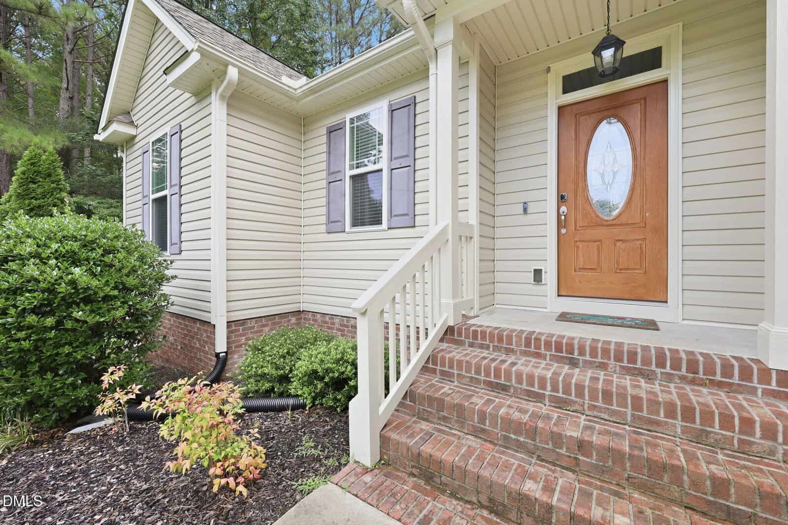 Front porch of a house with beige siding, a brick stairway, white railing, a wooden front door with glass panel, two shuttered windows, and landscape plants along the walkway.