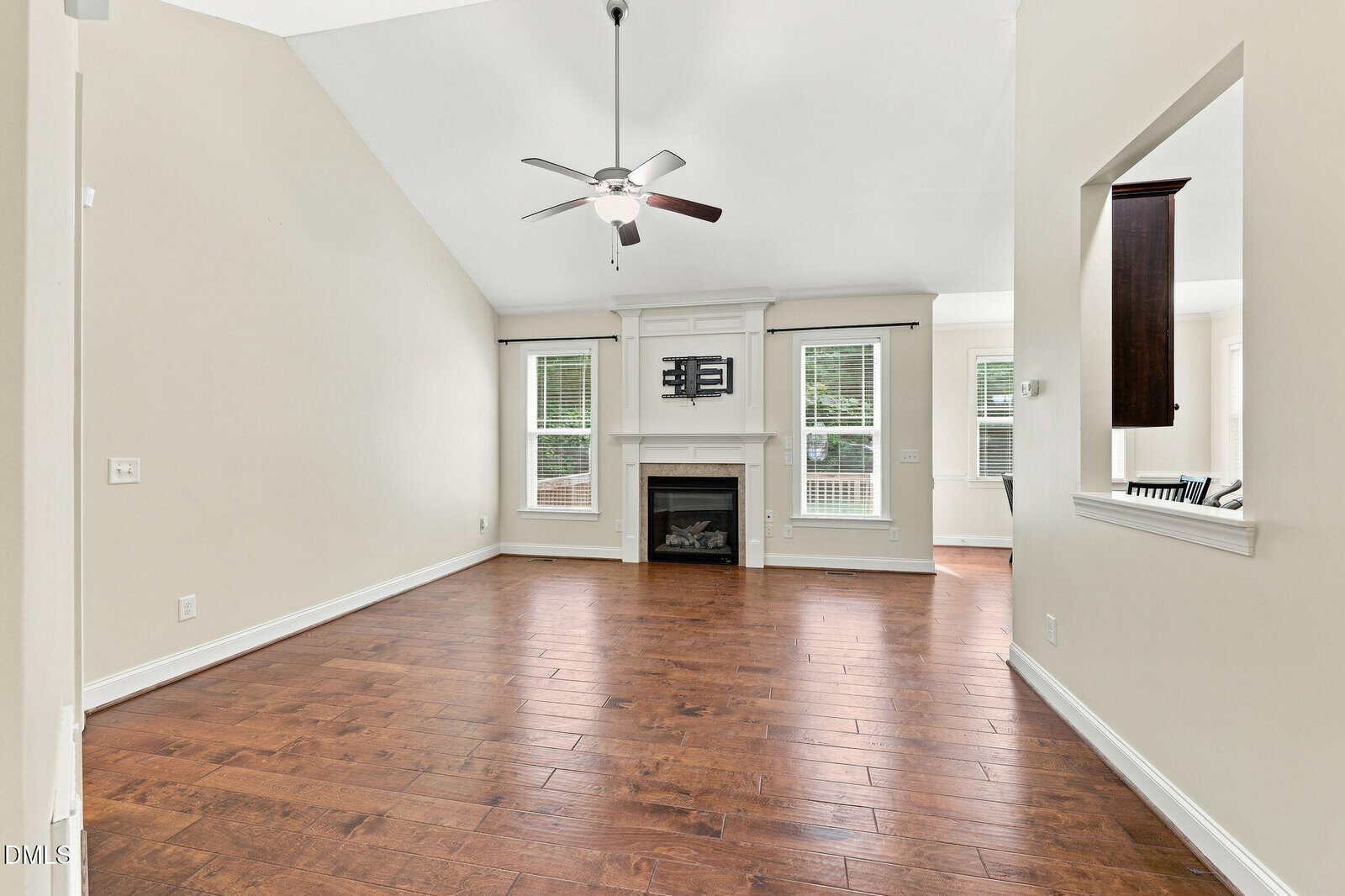 Spacious living room with hardwood floors, vaulted ceiling, ceiling fan, fireplace, mounted TV, two windows, and an open pass-through to the kitchen. Walls are light beige. Room is unfurnished.