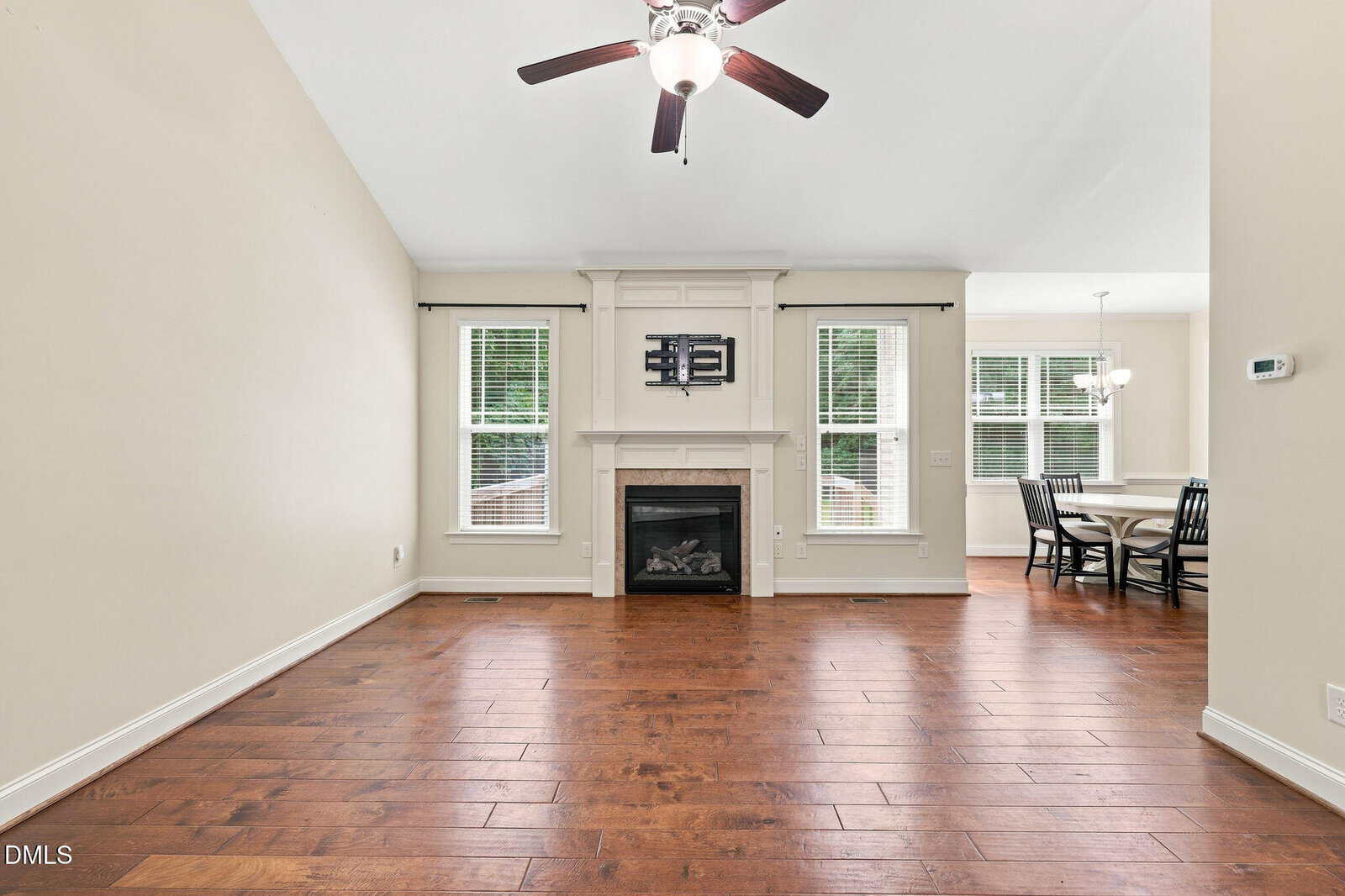 Unfurnished living room with hardwood floors, a ceiling fan, fireplace with a TV mount above, and two windows. Dining area with a round table and four chairs is visible to the right, next to more windows.