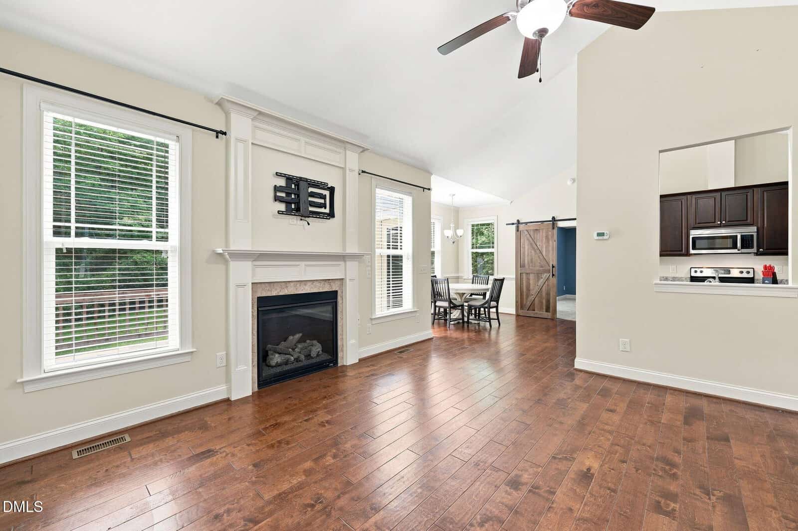 Bright living area with hardwood floors, a fireplace, wall-mounted TV, two tall windows, and a ceiling fan. Opens to a dining area with a farmhouse-style sliding door, and a kitchen visible through a pass-through window.