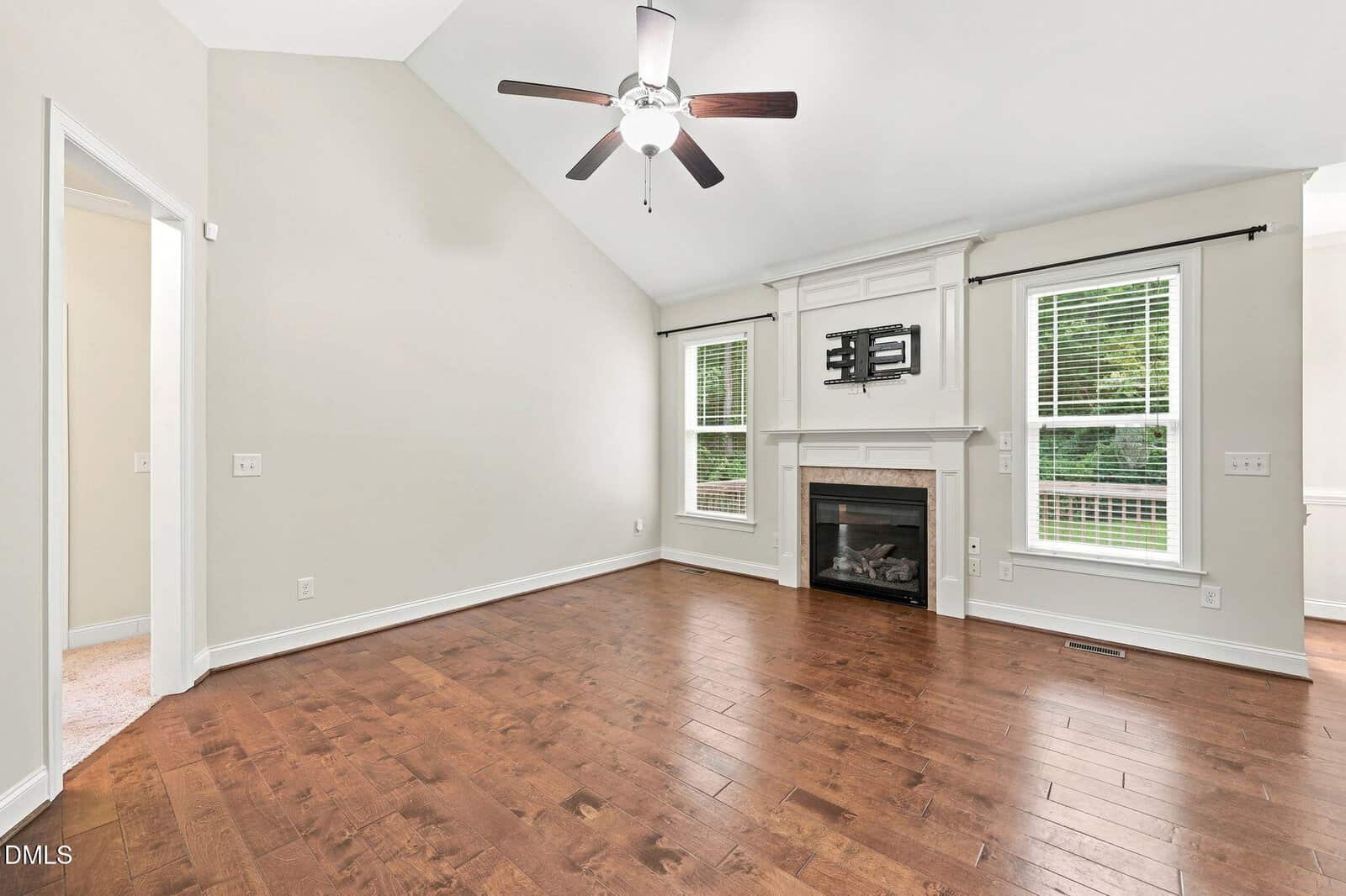 Bright living room with vaulted ceiling, ceiling fan, hardwood floors, two windows, a fireplace, and a wall mount for a TV above the mantle. Neutral-colored walls and natural light fill the space.