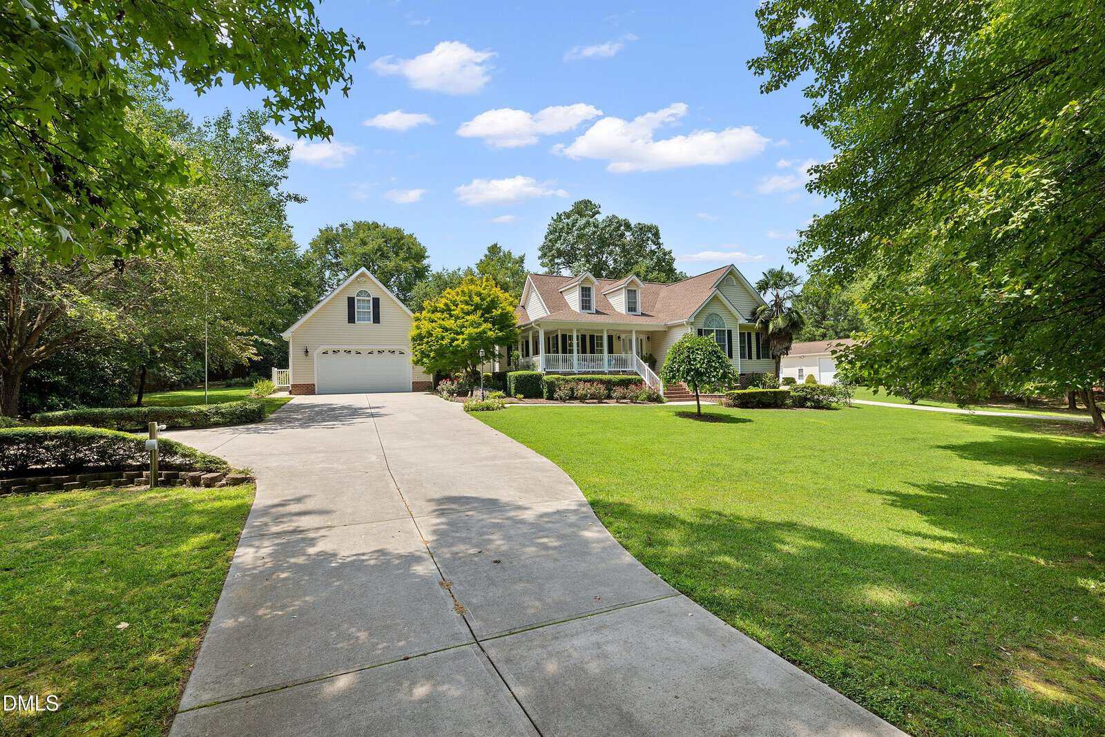 A two-story house with a large covered porch sits at the end of a curved driveway, surrounded by green grass, trees, and landscaped shrubs under a blue sky with scattered clouds.