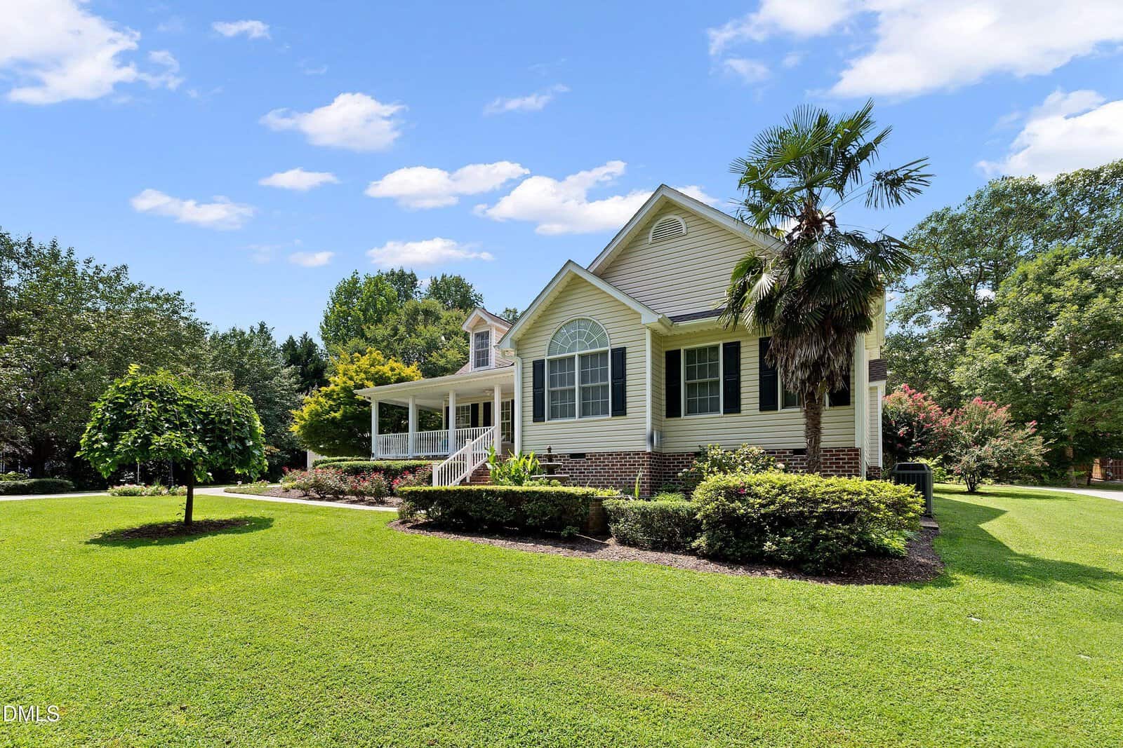 Single-story beige house with white trim, large windows, a covered front porch, and neatly trimmed landscaping. The yard is spacious and green, with mature trees and bushes. The sky is mostly clear with scattered clouds.