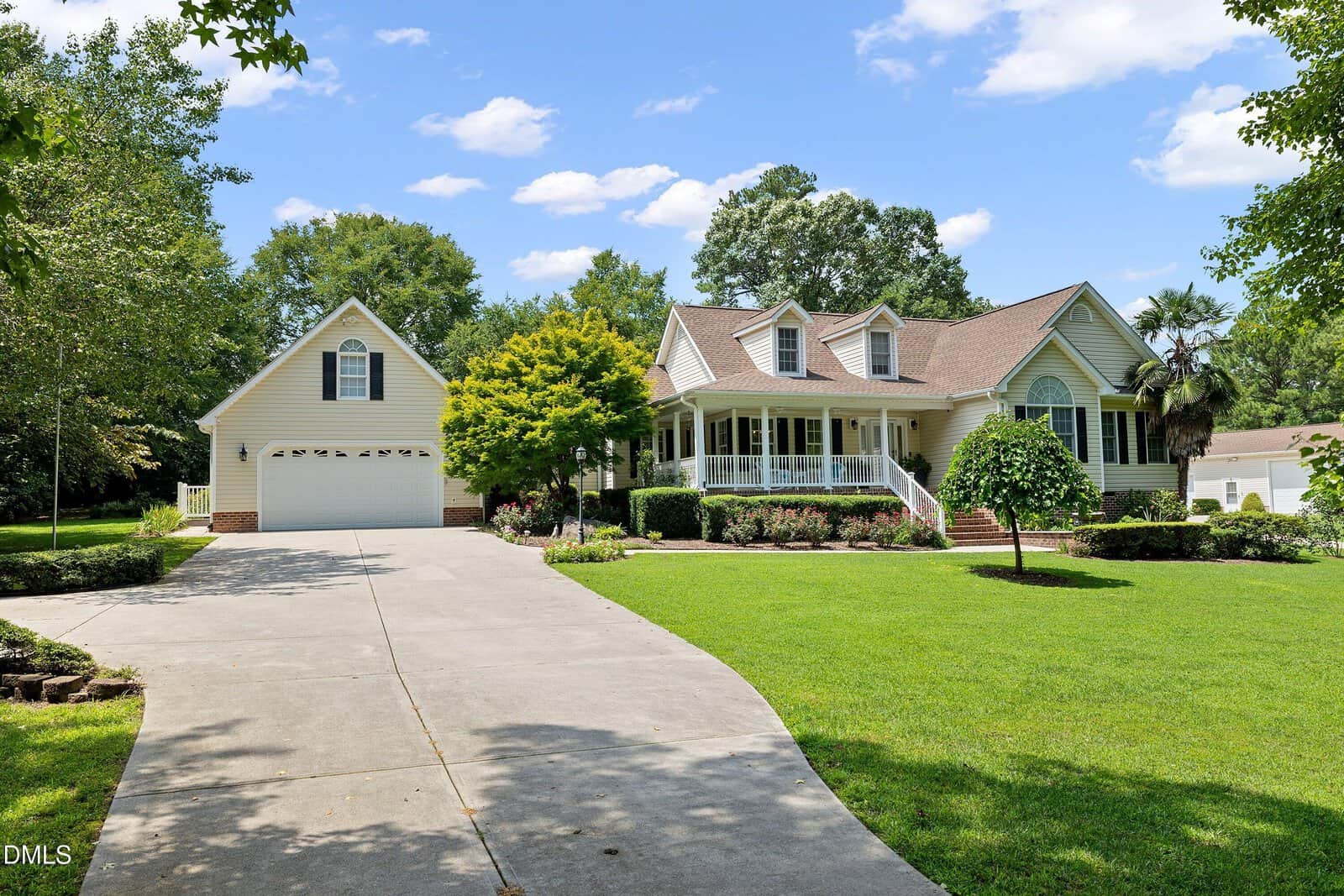 A large suburban house with beige siding, a covered front porch, dormer windows, an attached garage, and a wide driveway sits at 15 Winter Woods Drive in Louisburg. The yard is landscaped with grass, trees, and shrubs under a partly cloudy sky.